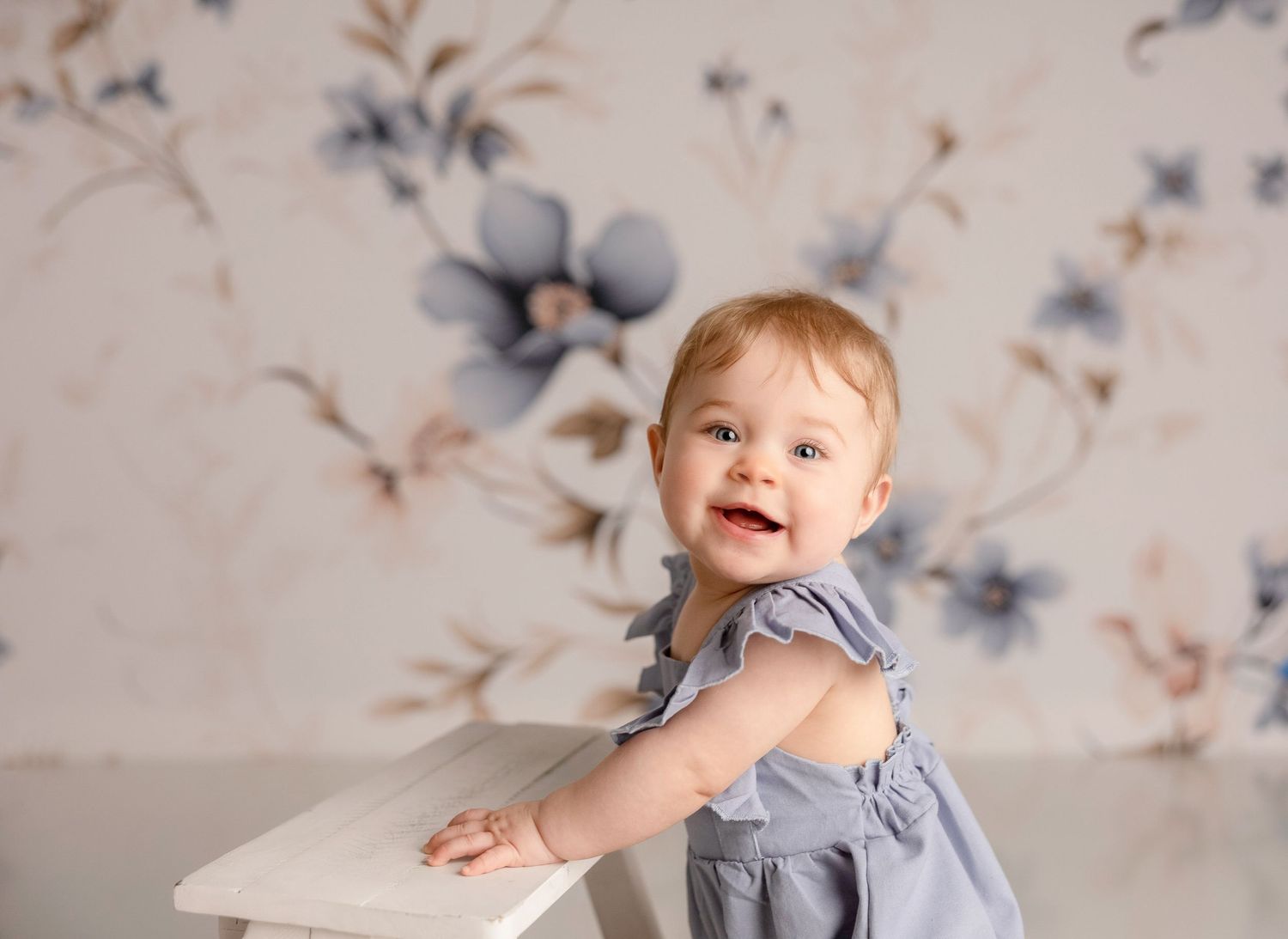 Joyful baby in blue dress against floral wallpaper background.