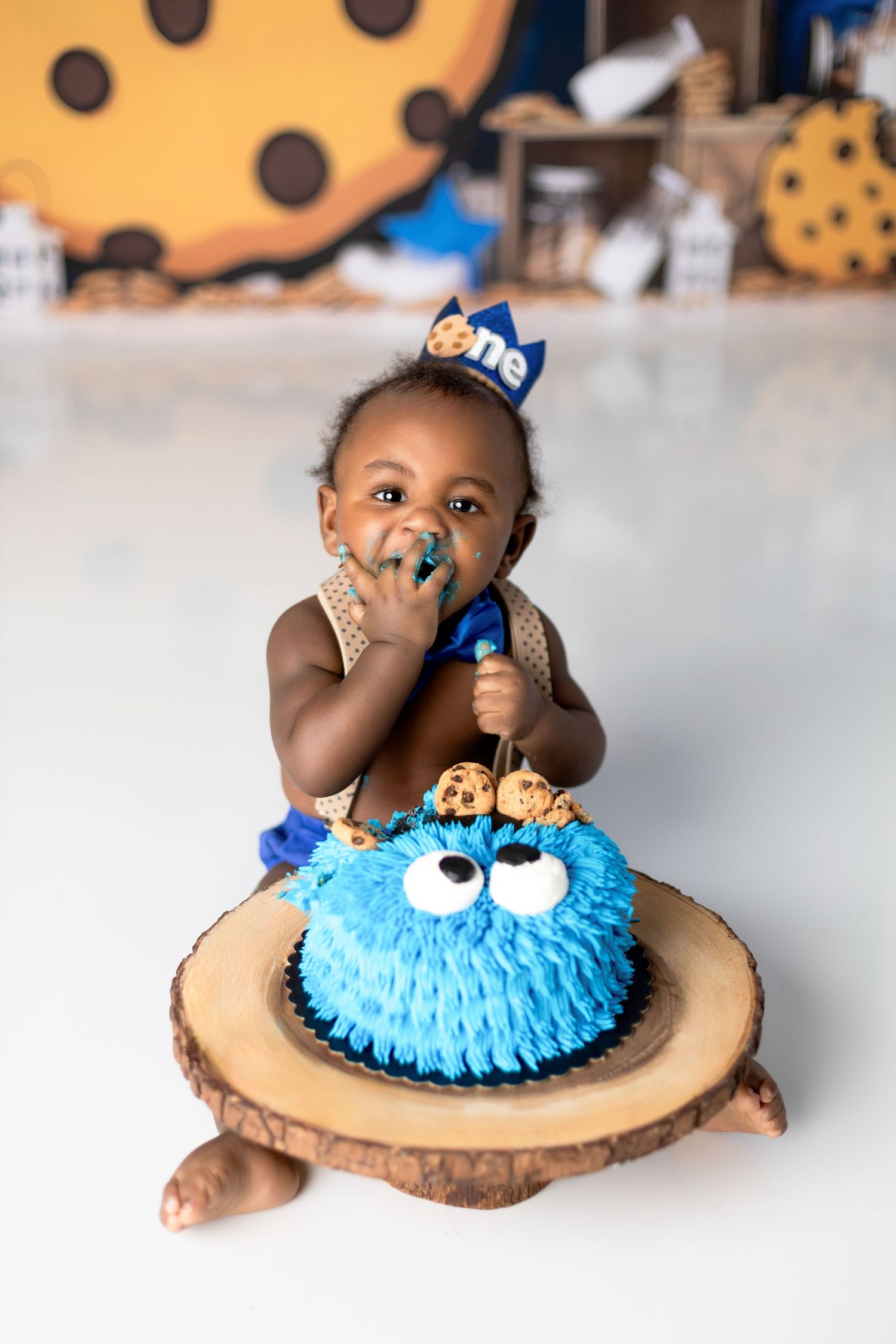 Toddler in blue crown sitting with Cookie Monster cake on wooden slice platform.