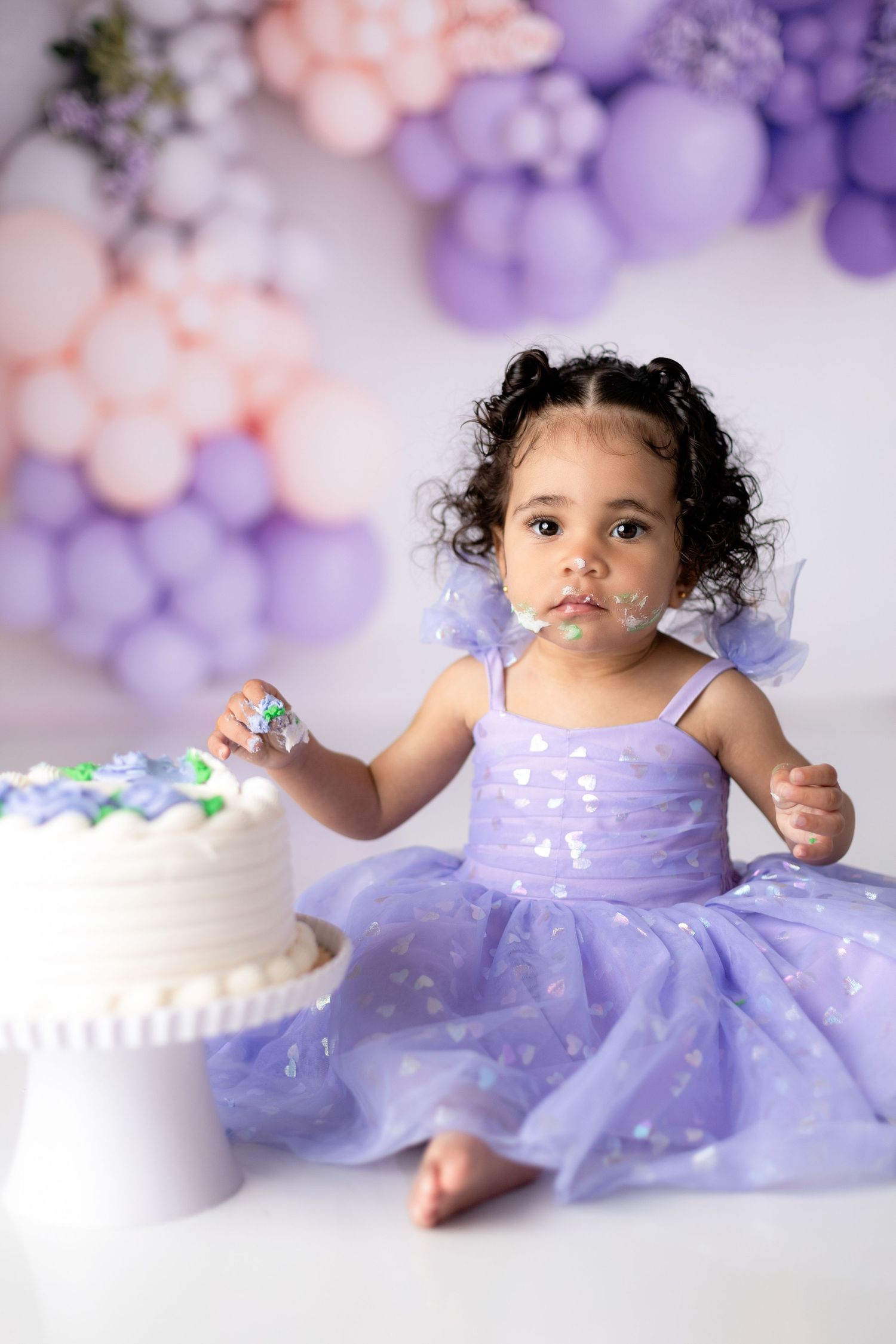 Cake smash photo of toddler in purple tulle dress with balloon backdrop.