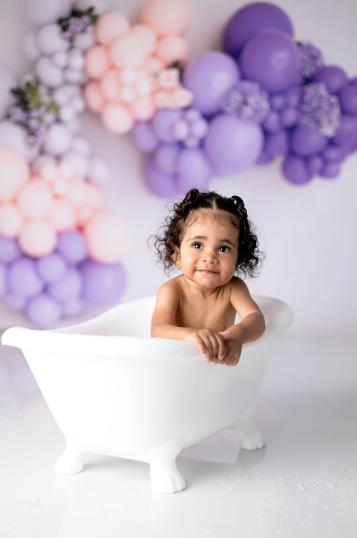 Toddler posing in miniature white bathtub with purple and pink balloon decorations.