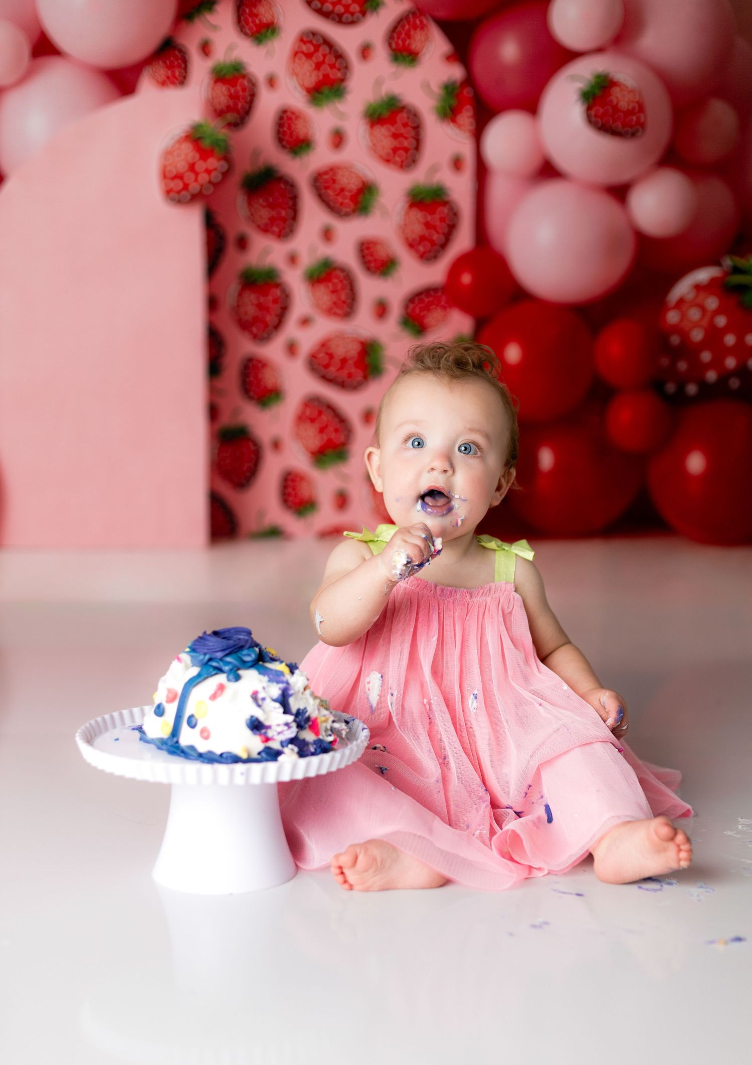 Baby in pink outfit enjoys cake smash photoshoot with strawberry themed decorations.