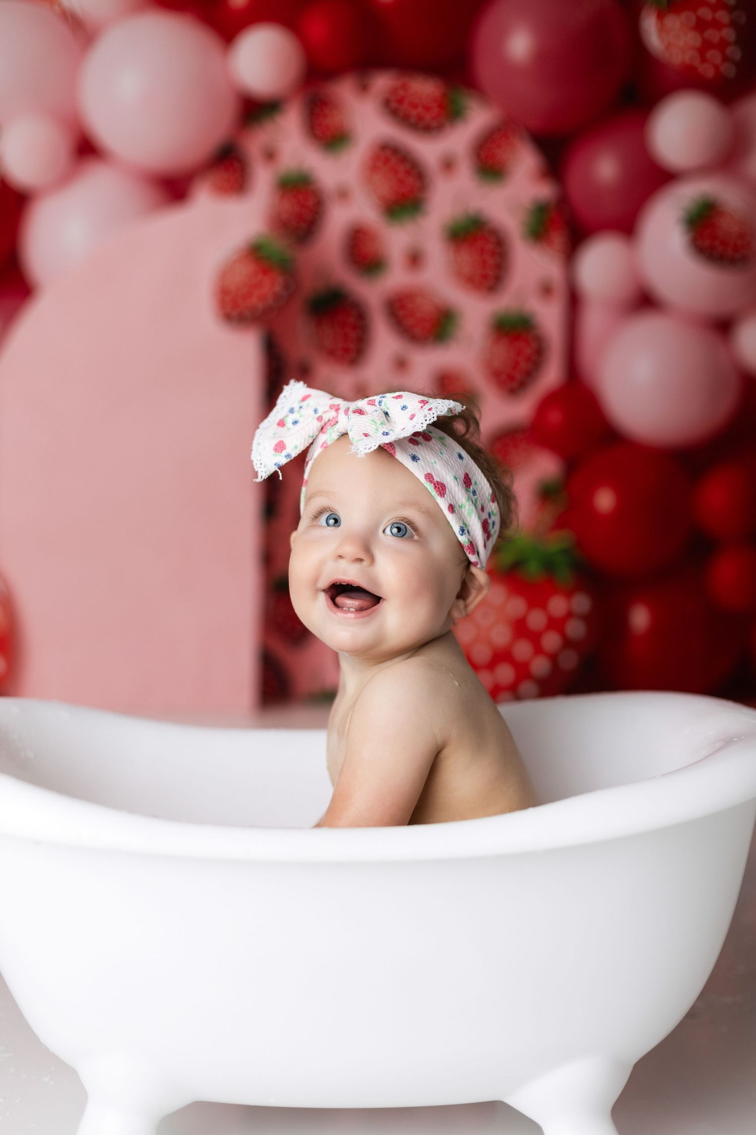 Baby in strawberry-themed photoshoot sits in white bathtub with pink balloon backdrop.