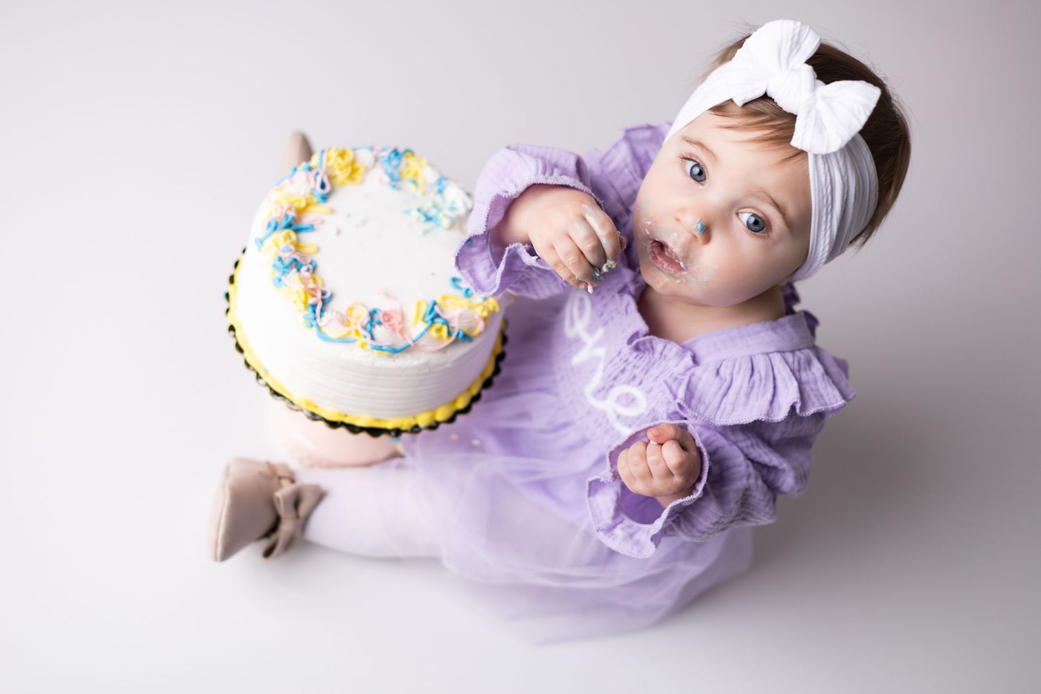 Baby in purple tulle dress lying next to decorated birthday cake on white backdrop.