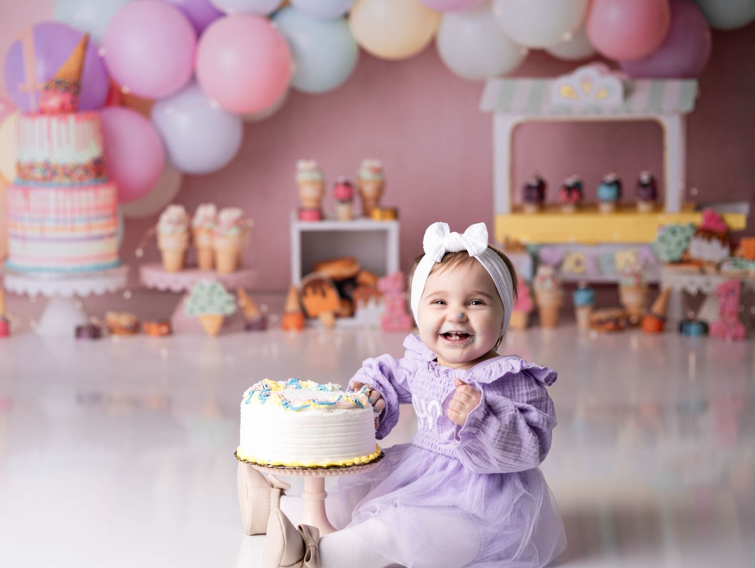 First birthday setup with pastel balloons, cake and decorations against pink backdrop.
