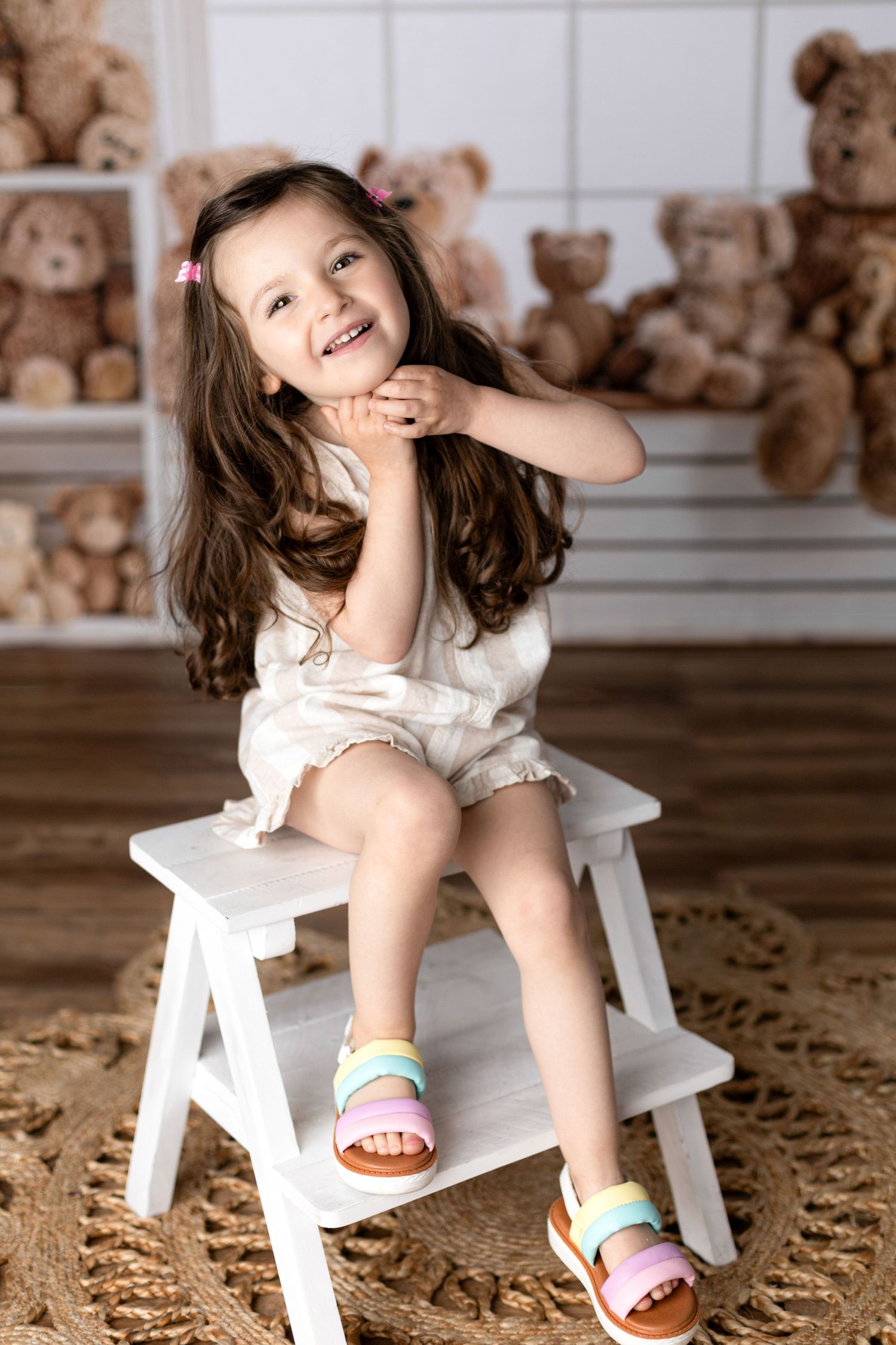 Young child in white outfit sits on white stool surrounded by teddy bears.