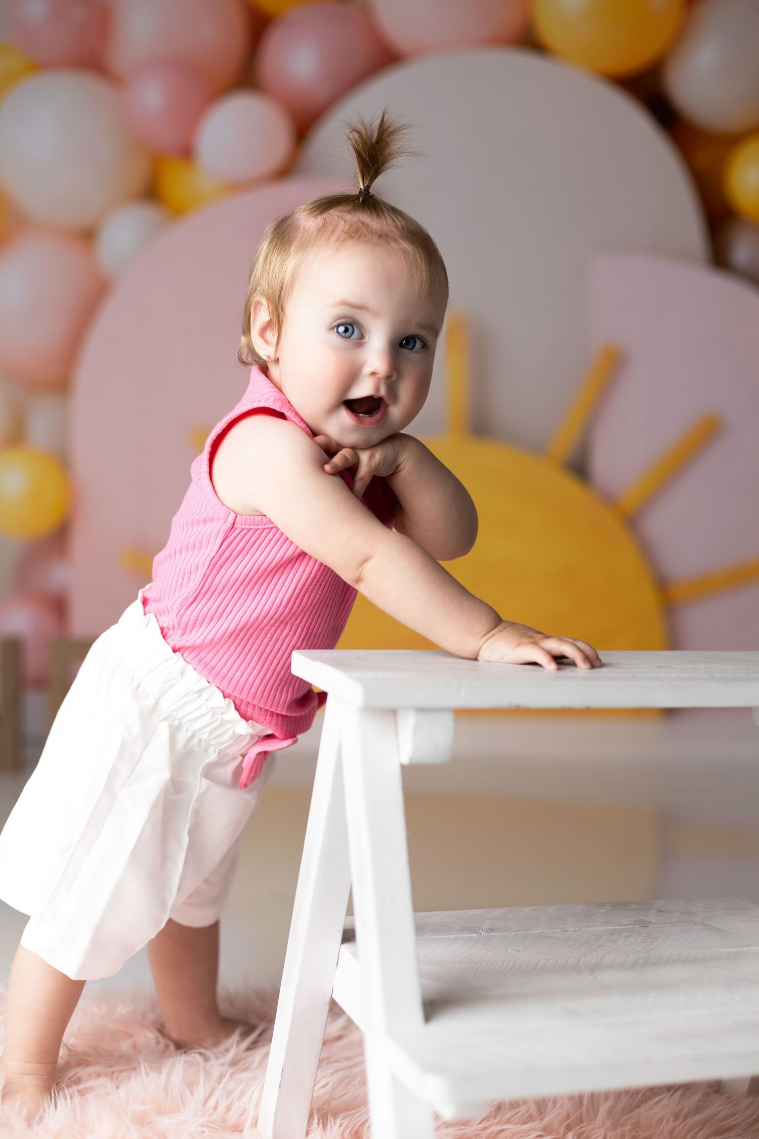 Excited baby in pink and white outfit leans on white bench with pastel balloon backdrop.