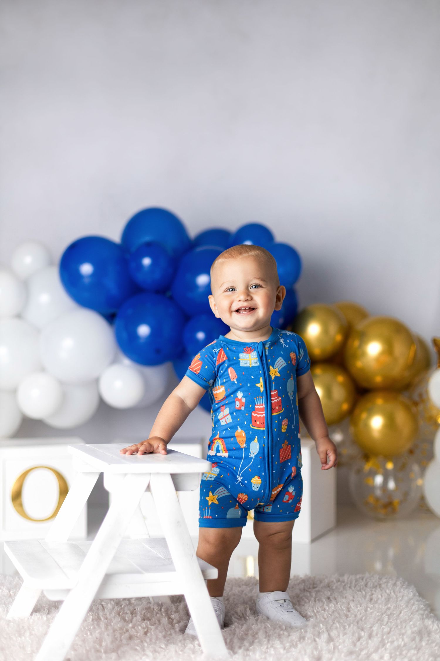 Toddler in blue patterned romper stands by white bench with blue, white and gold balloon backdrop.