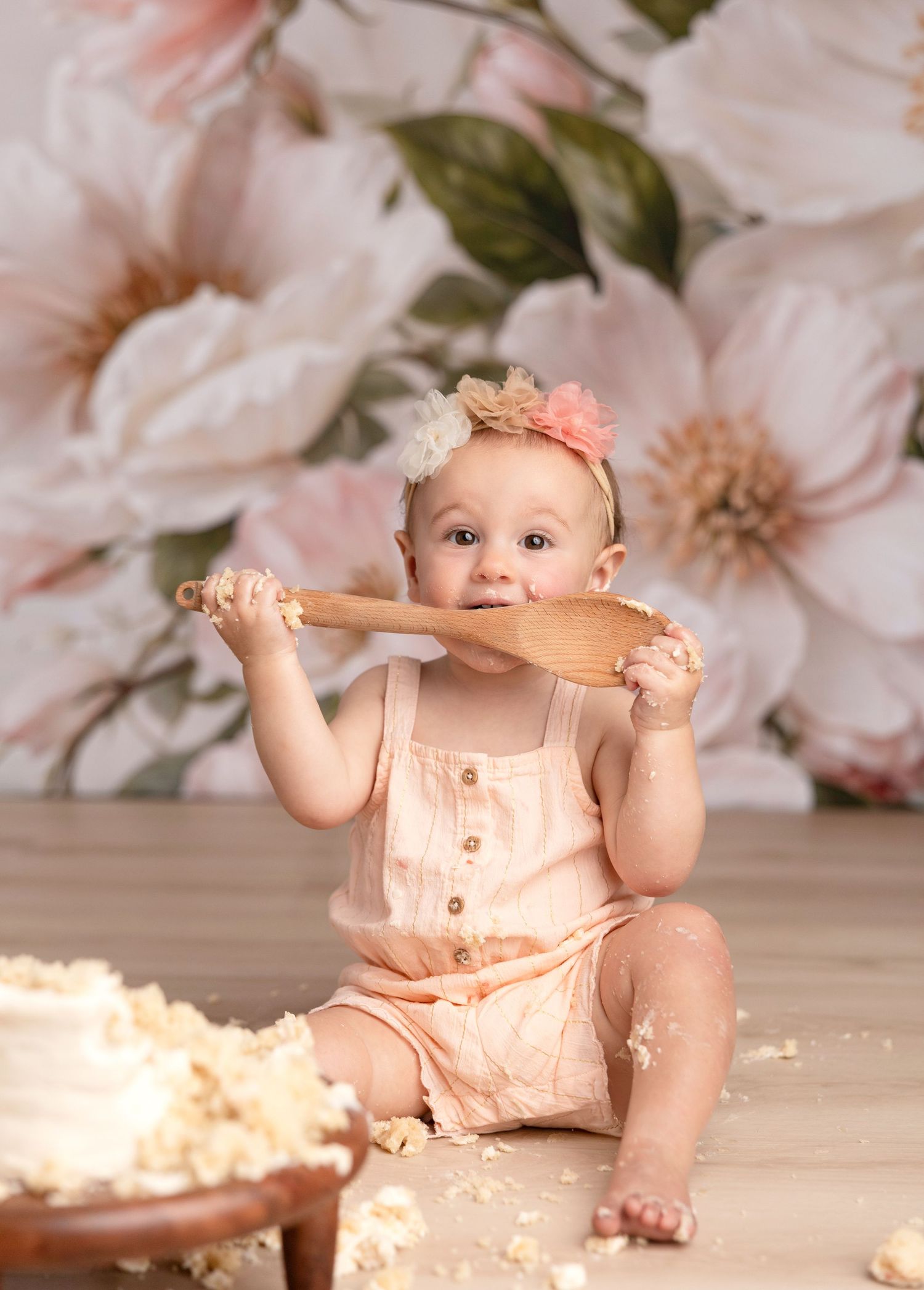 Baby in peach romper playfully bites wooden spoon during cake smash photo session with floral backdrop.