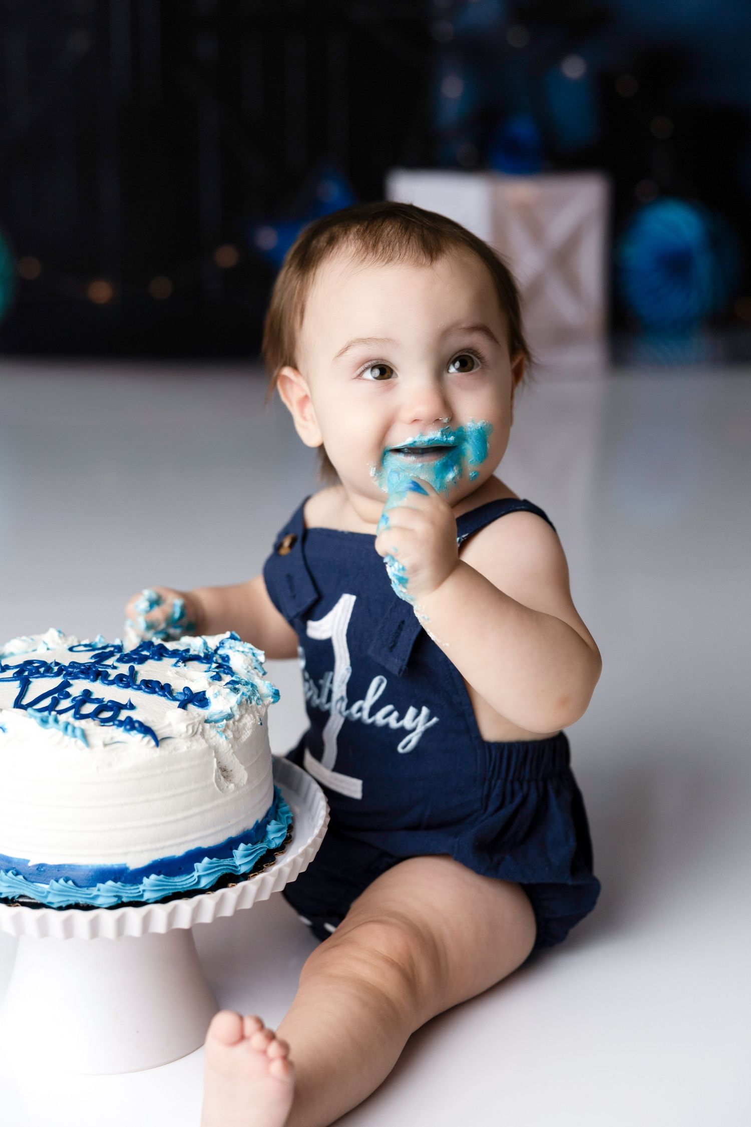 Baby in navy blue outfit sits next to white birthday cake with blue frosting decorations.