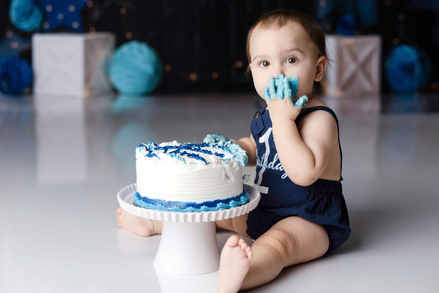 Baby with blue pacifier sits beside white and blue decorated cake on cake stand.