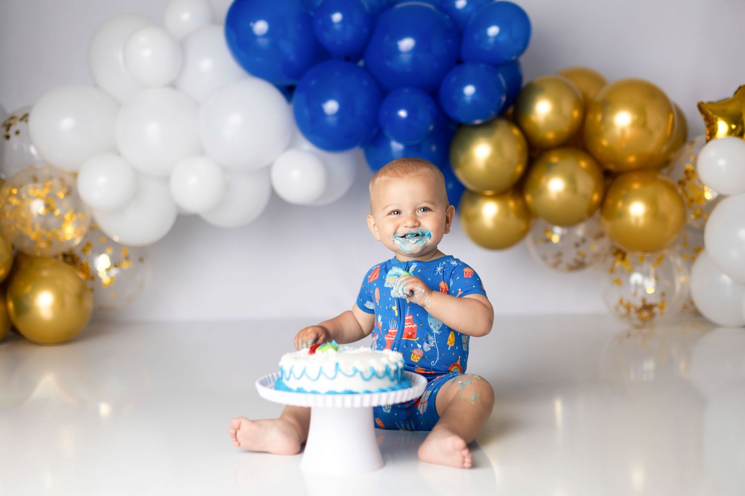 Baby in blue outfit sits with birthday cake surrounded by festive balloons.
