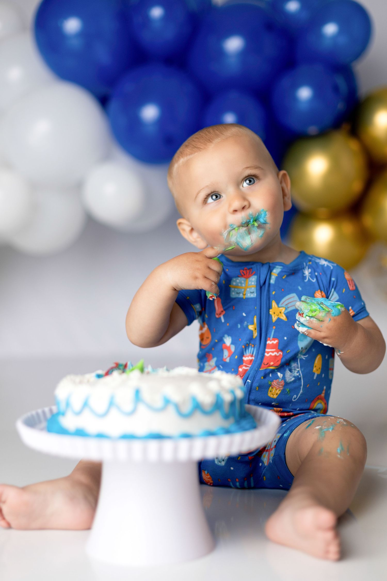 Baby with frosting-covered face enjoys smashing birthday cake with blue and gold balloon decor.
