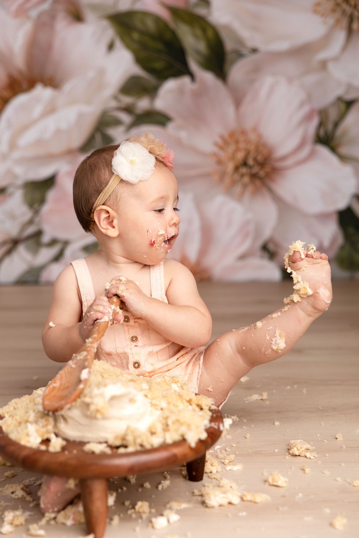 Baby in peach outfit enjoys messy cake smash photoshoot against floral background.
