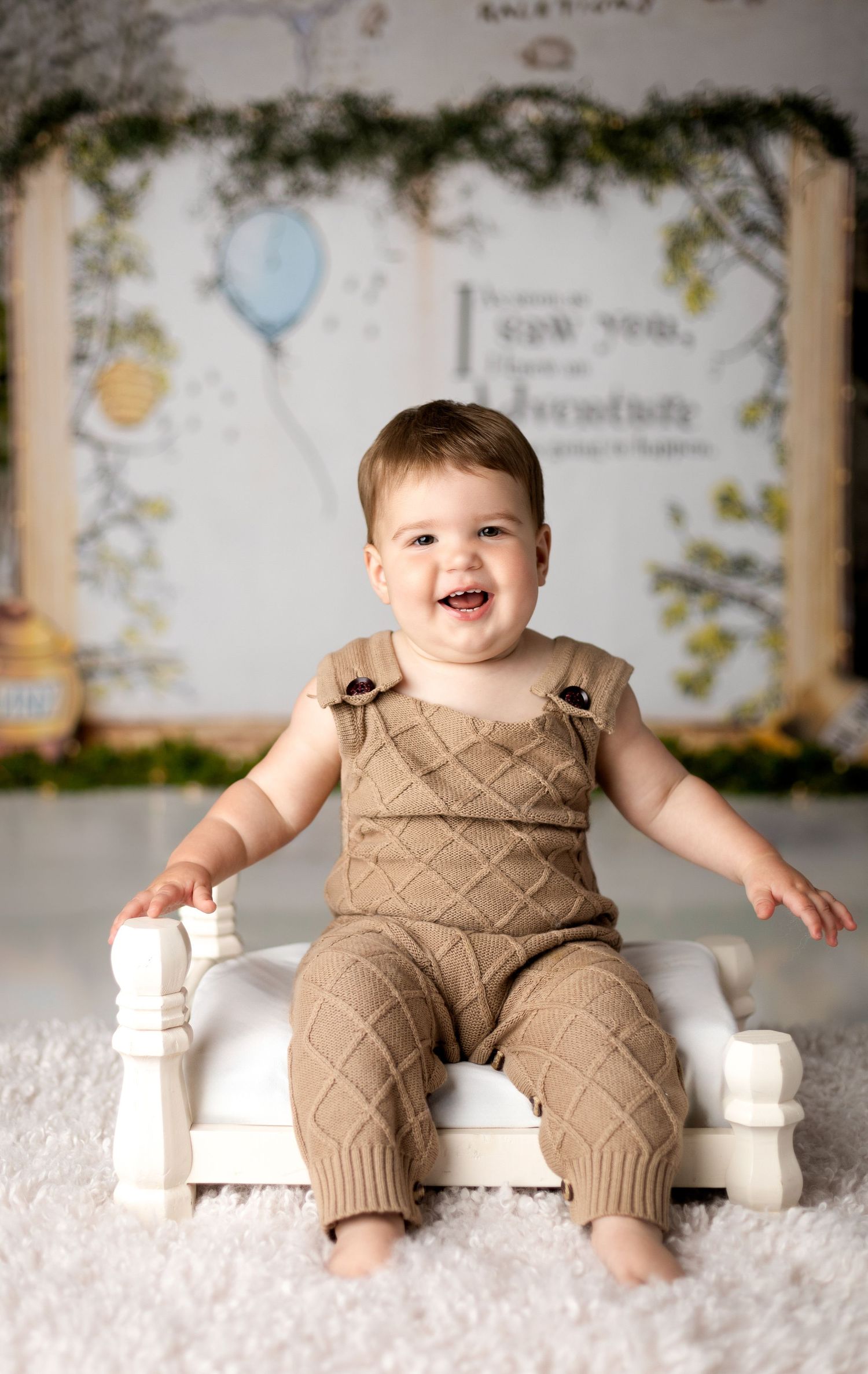 Happy baby in tan quilted romper sits on white bench with decorated backdrop.