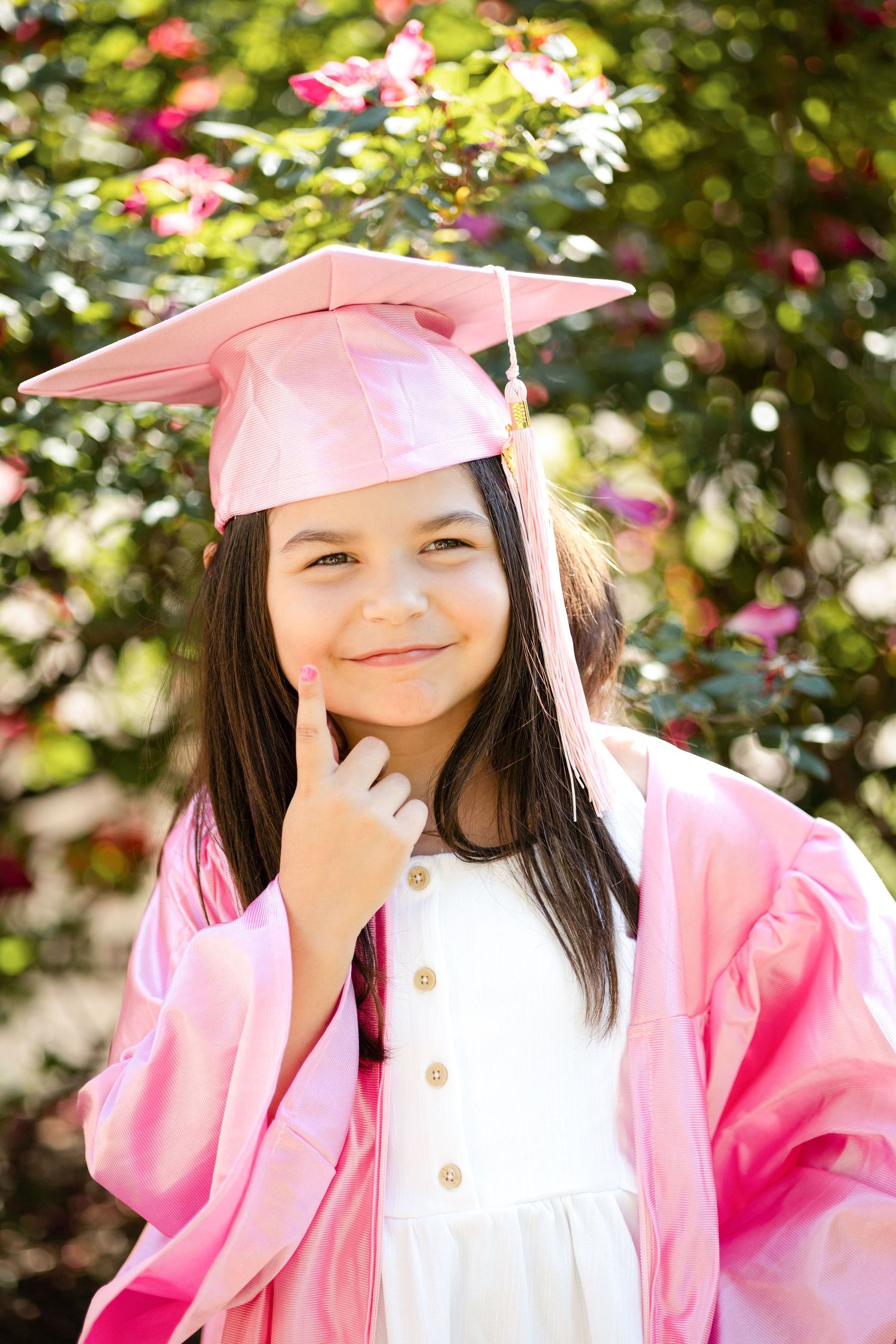 Young graduate in pink cap and gown smiling outdoors with floral background.