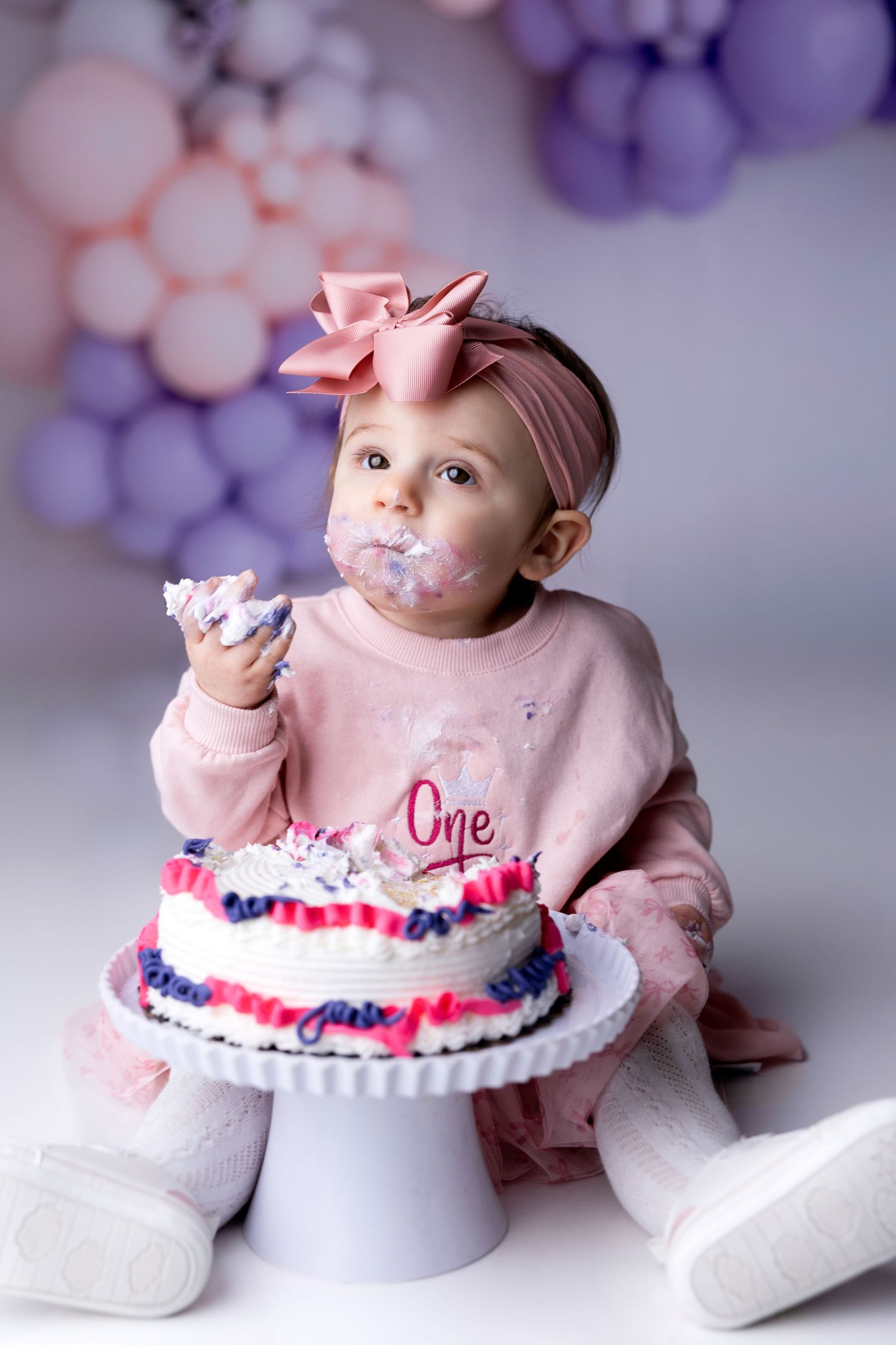 Baby in pink outfit with bow headband enjoys first birthday cake smash photoshoot.