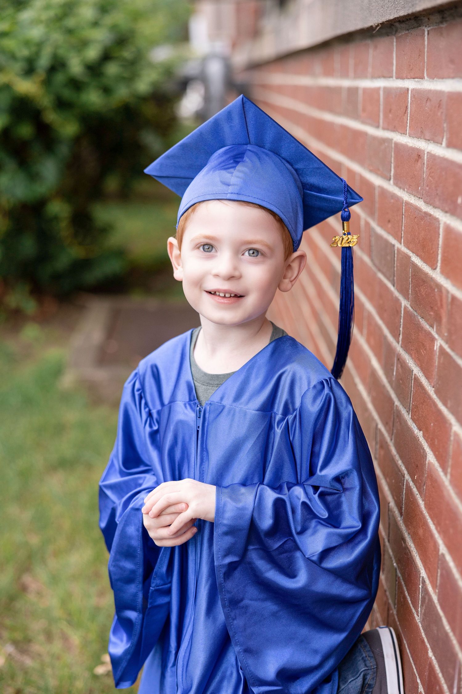 Young graduate in bright blue cap and gown sitting against brick wall outdoors.