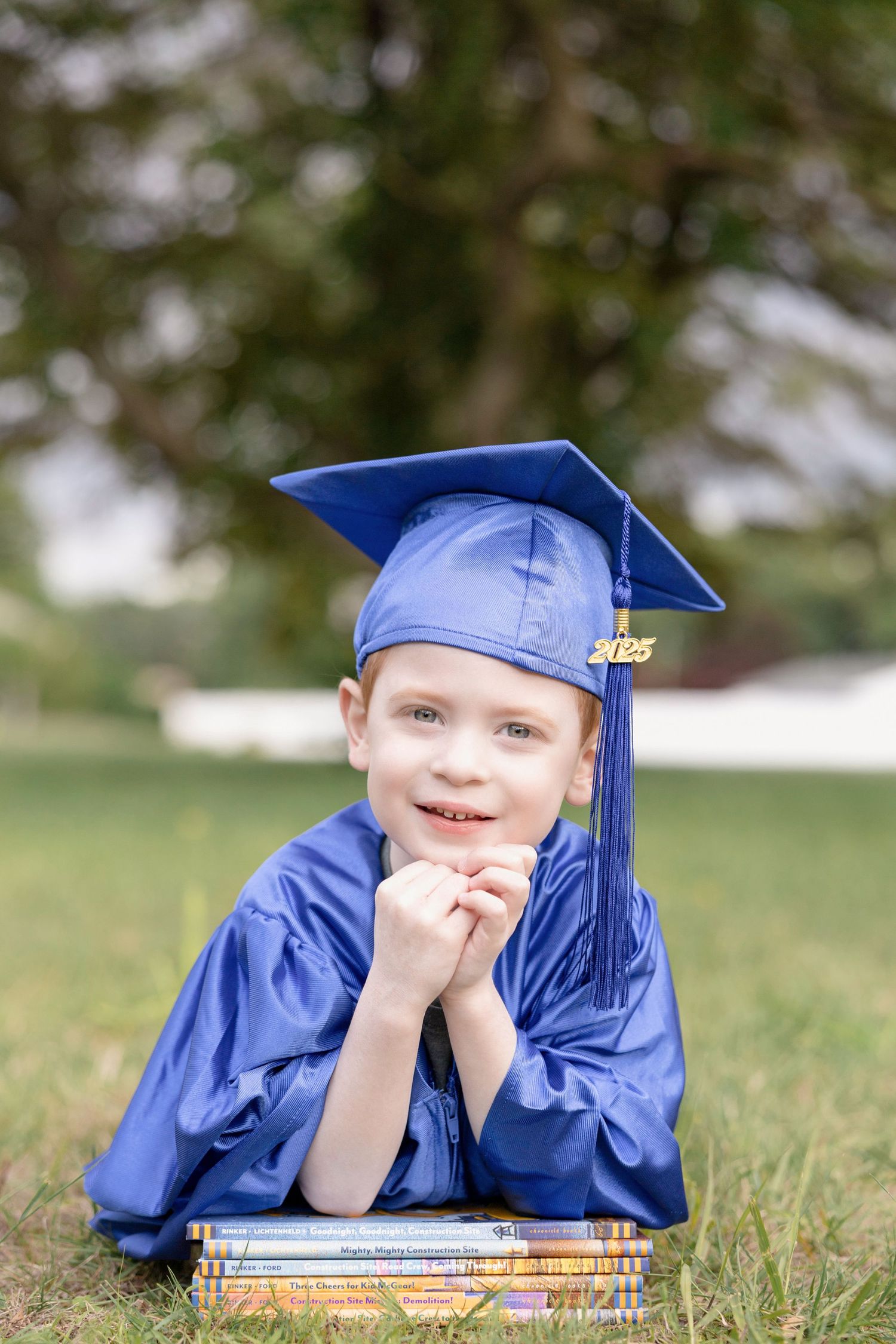Graduate in blue cap and gown lying on grass with books.