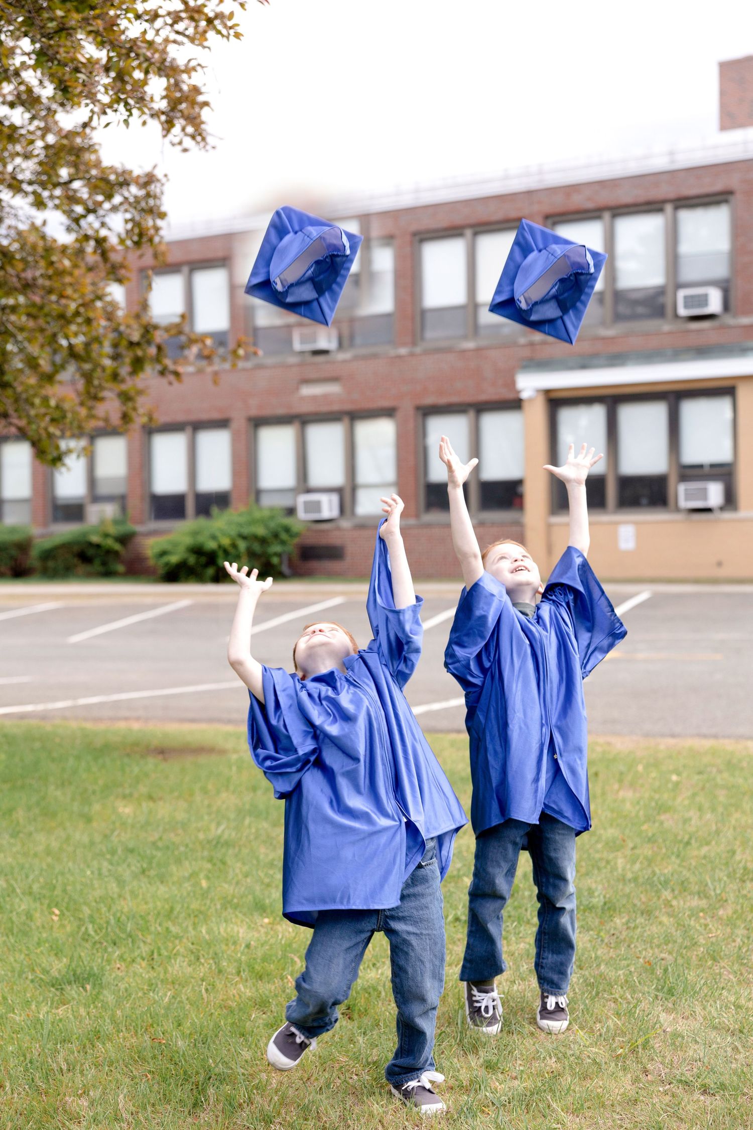 Children in blue graduation gowns running and jumping on grass near brick building.