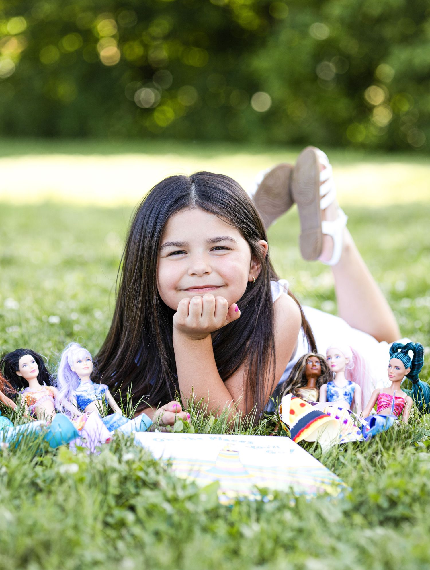 Young person lying on grass with collection of dolls and toys during outdoor photoshoot.