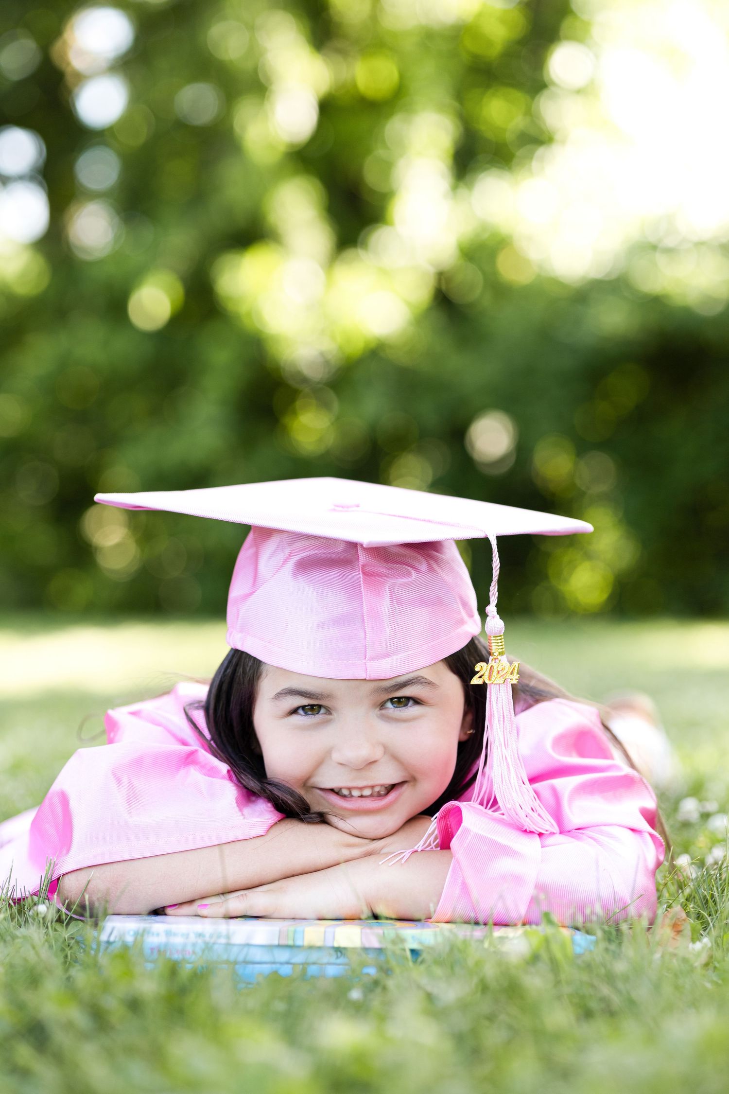 Graduate in pink cap and gown lying on grass during outdoor portrait session.