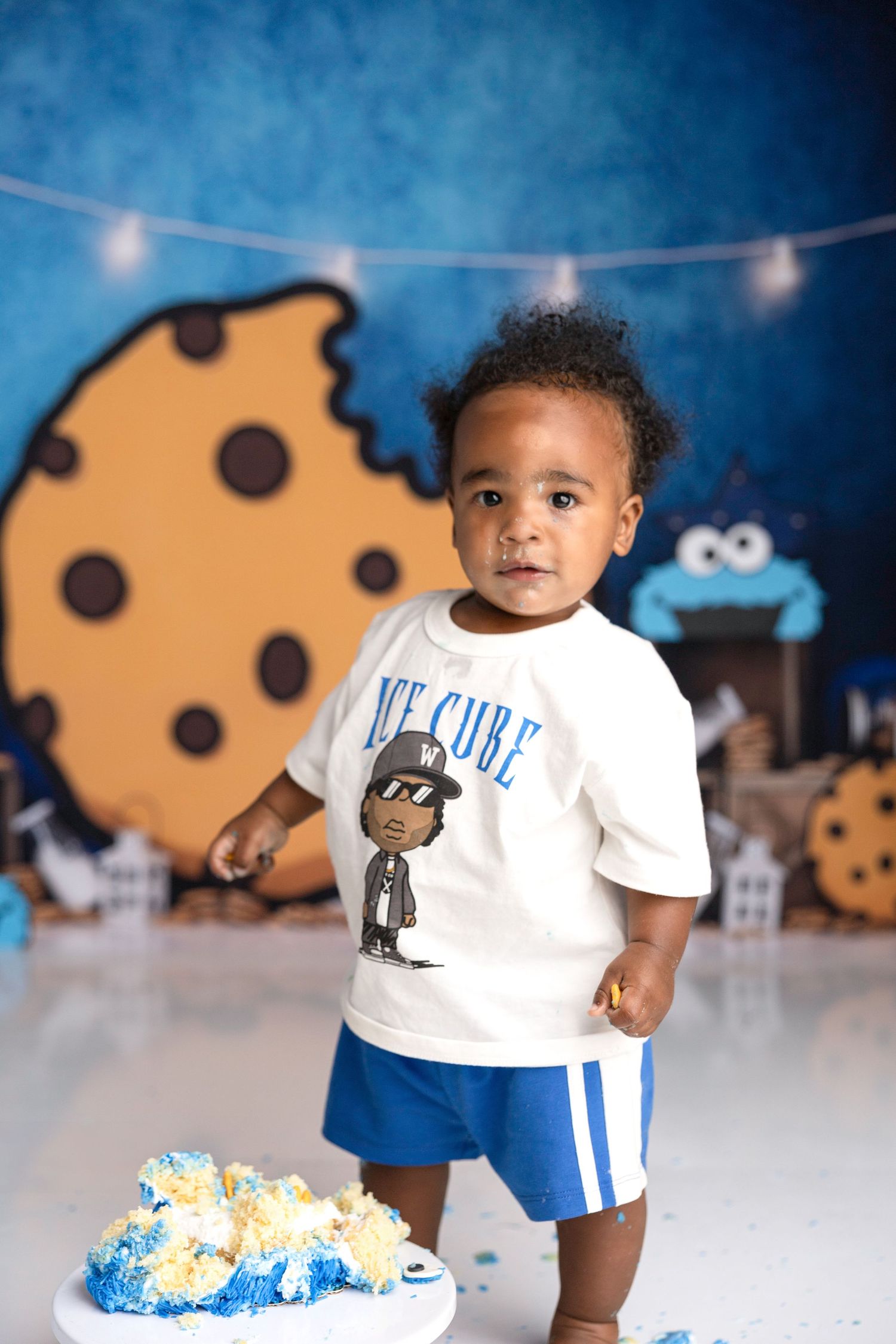 Baby in white t-shirt and blue shorts standing against cookie-themed backdrop.