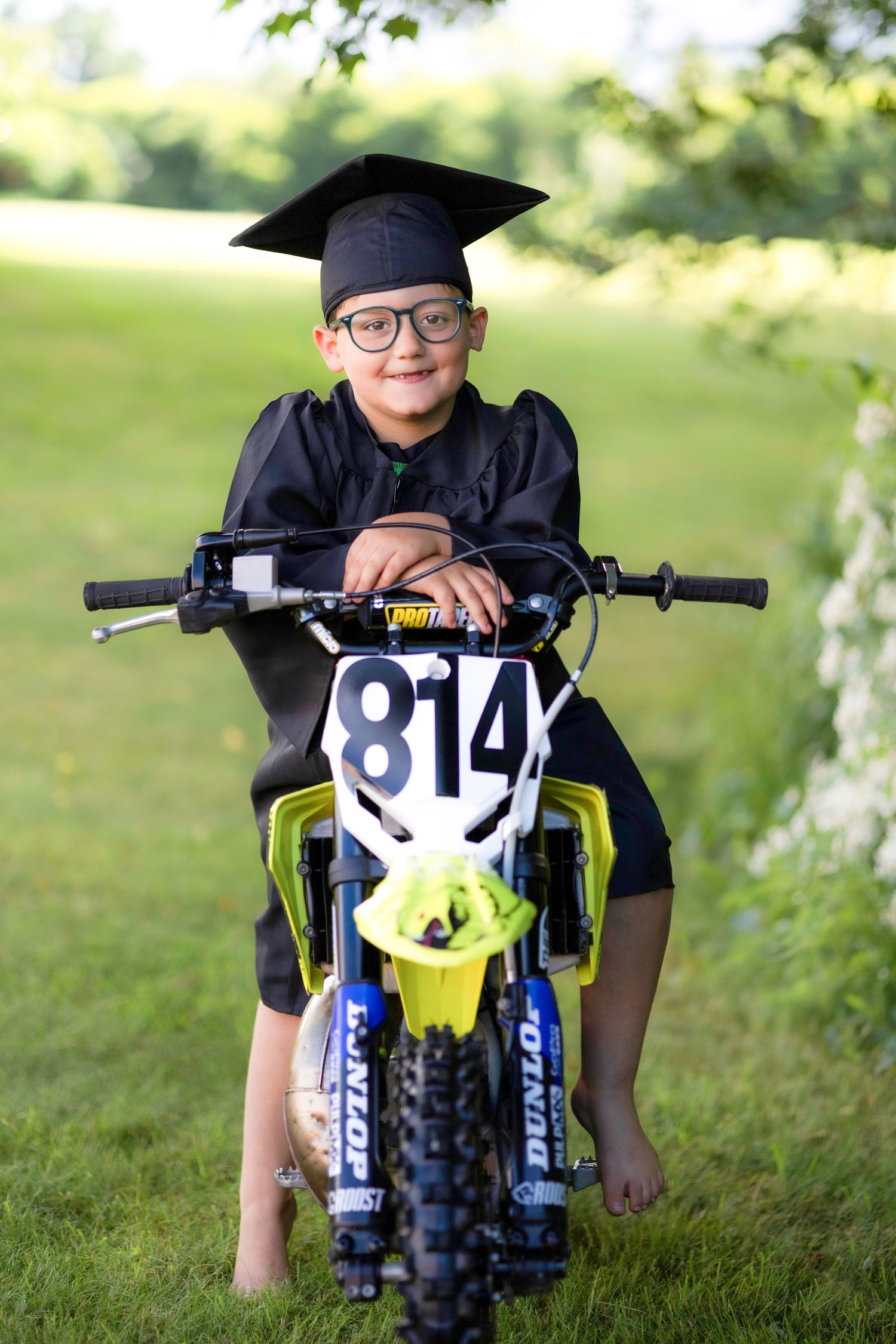 Young graduate in cap and gown sitting on dirt bike outdoors.