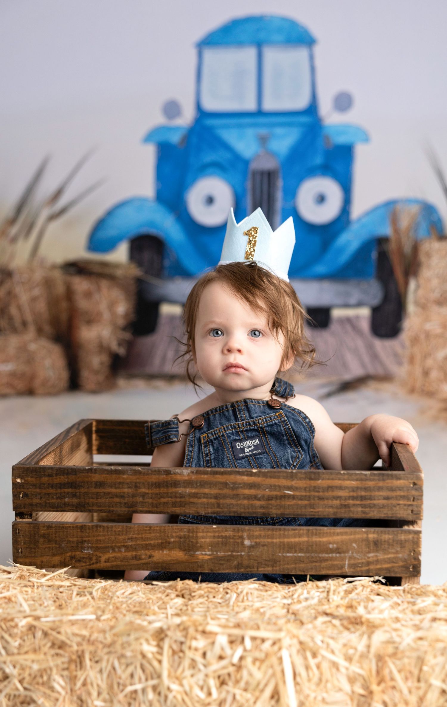 Baby sitting in rustic wooden crate with blue truck backdrop and hay bales.