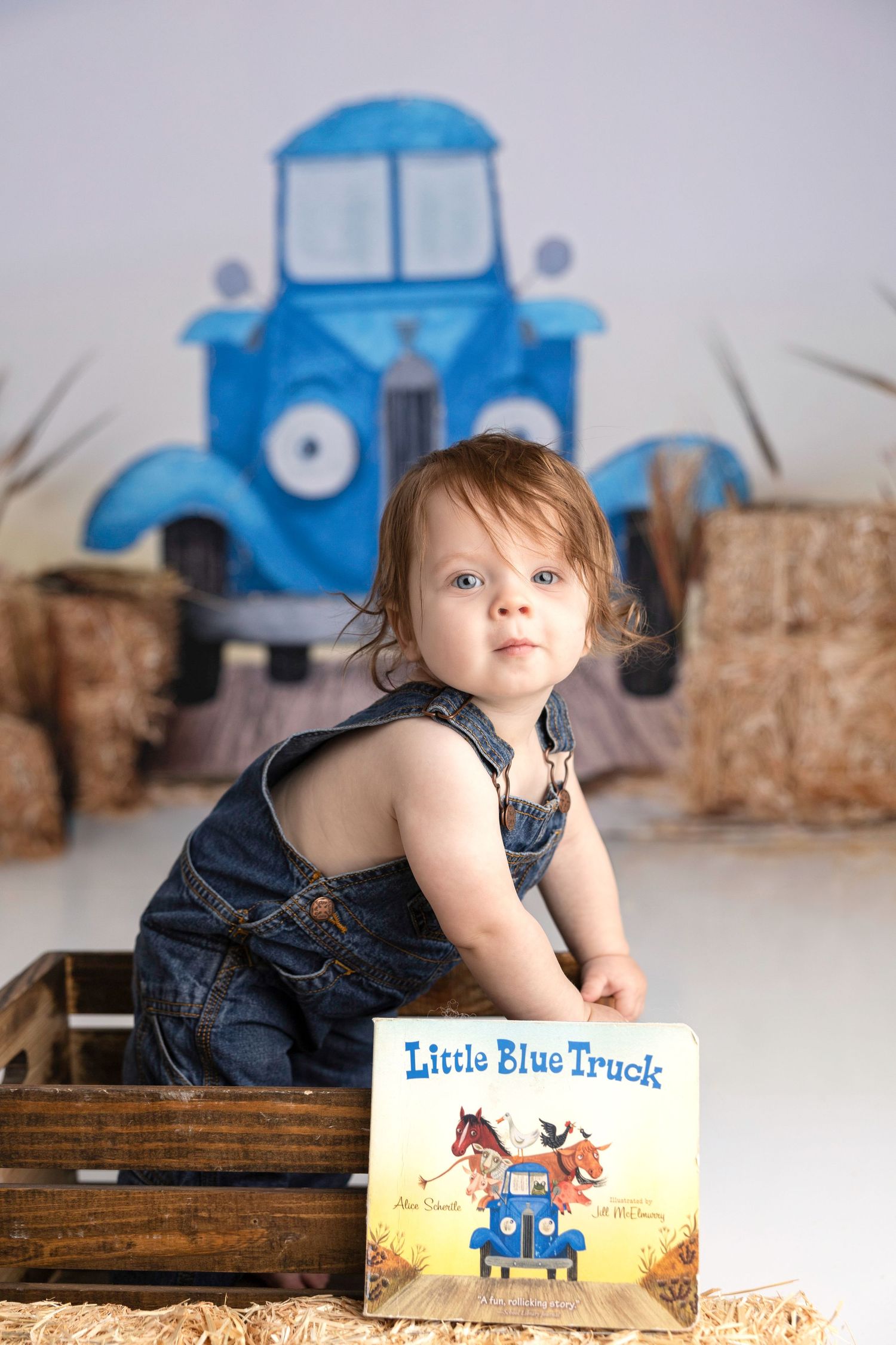 Baby in denim overalls posing with Little Blue Truck book against truck backdrop.