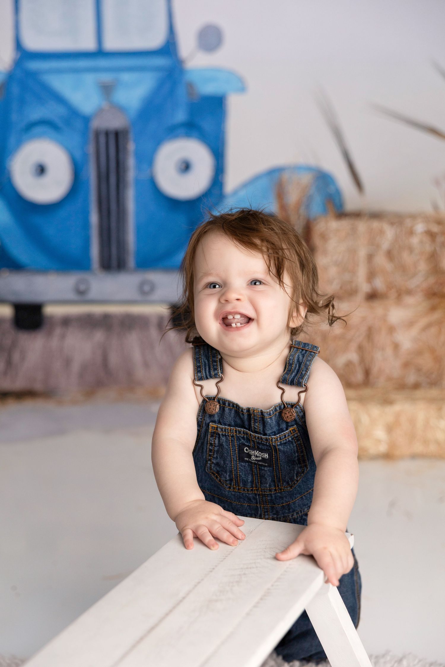 Baby in denim overalls laughing while sitting against blue truck backdrop.