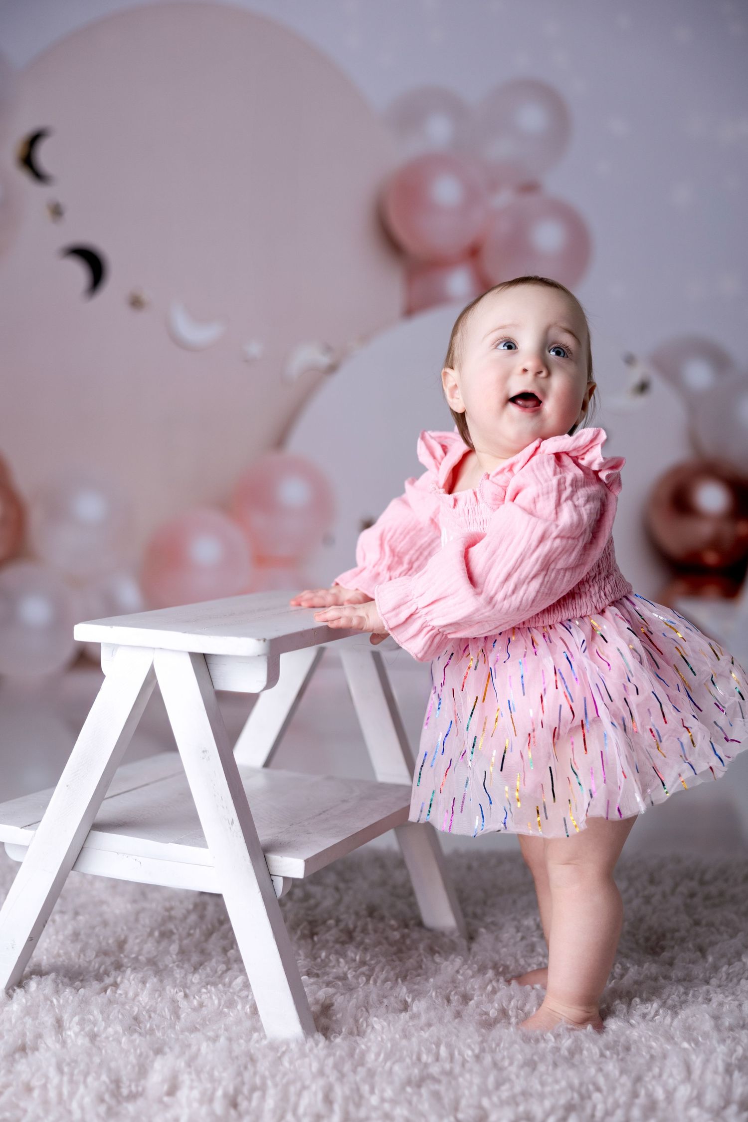 Small child in pink tulle dress stands next to white stool with balloon decorations.