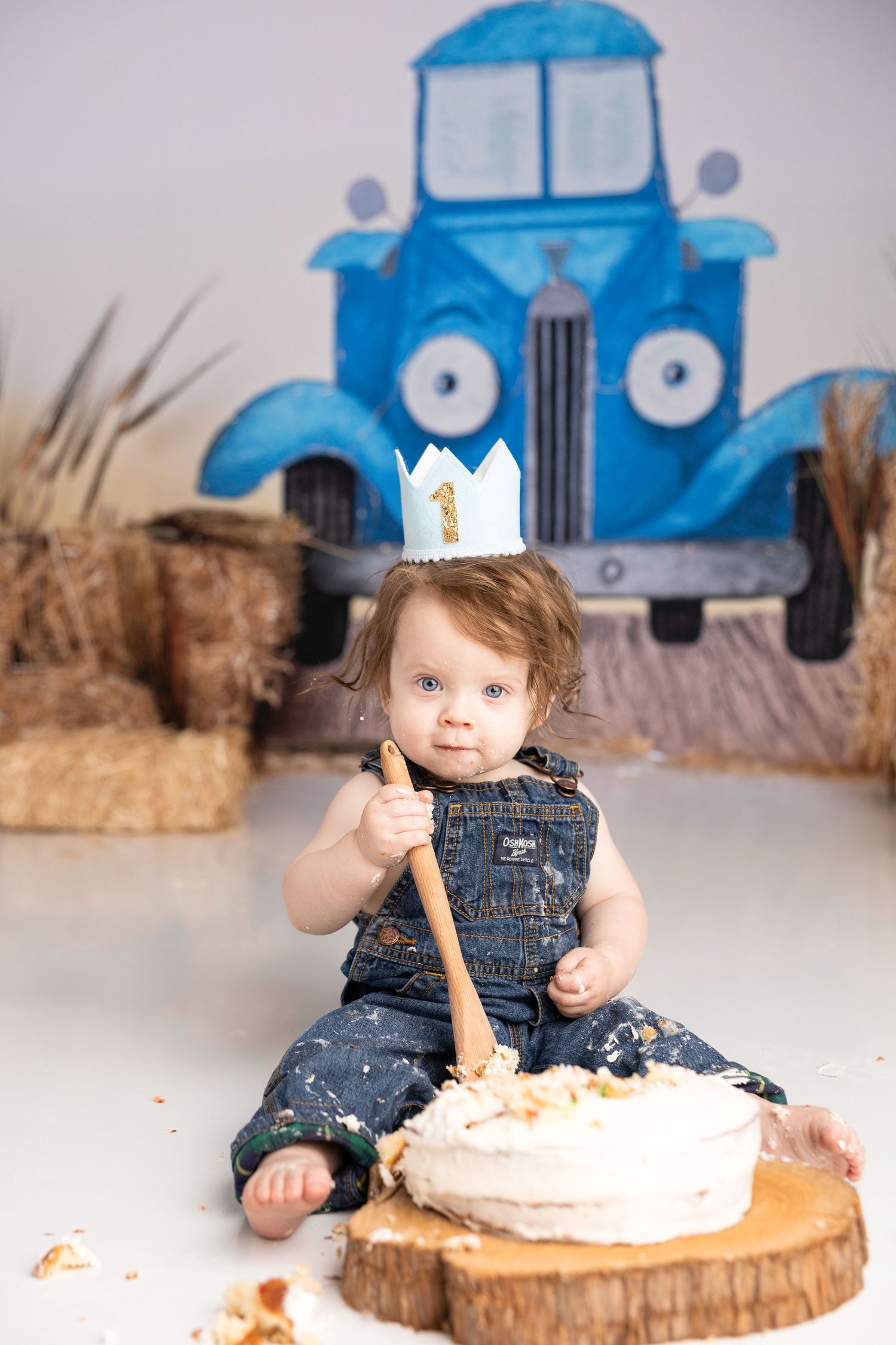 Baby in denim overalls enjoying cake smash on wooden slice with blue truck backdrop.