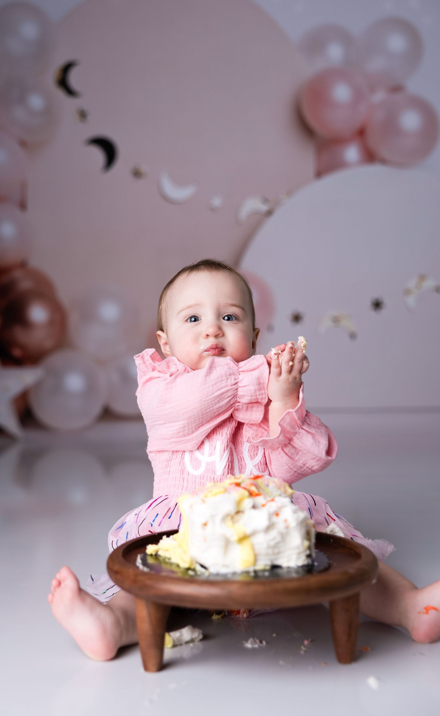 Baby in pink dress sits at wooden table with cake during birthday photoshoot.