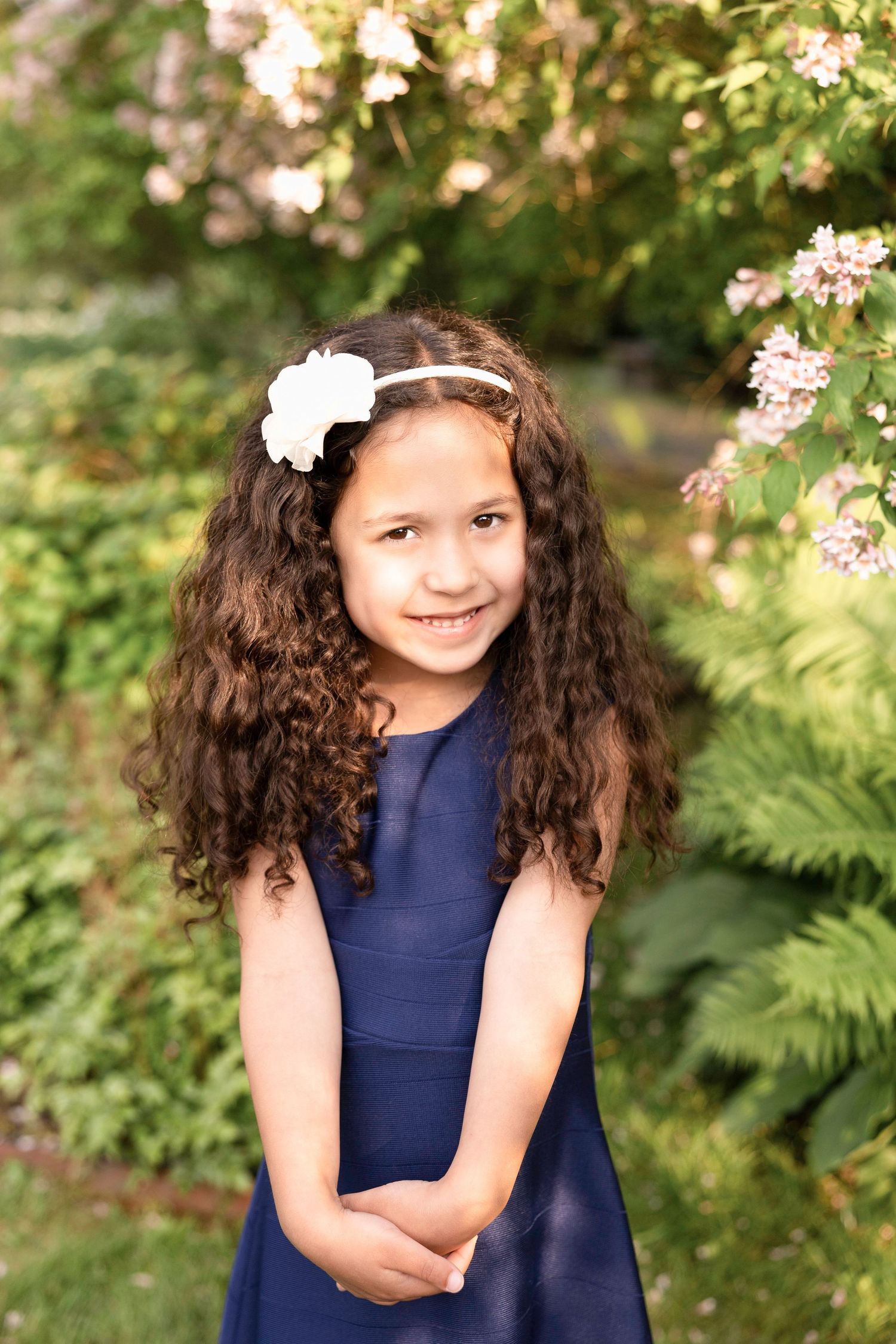 Young girl in navy dress with curly hair and white flower accessories in garden setting.