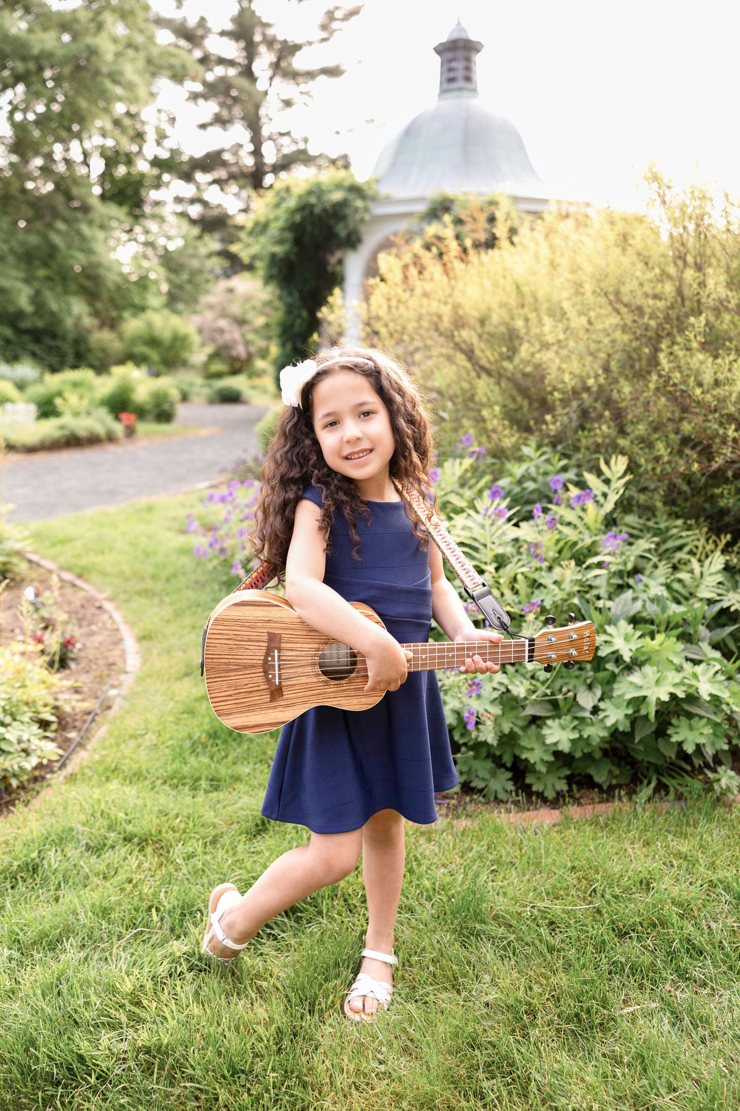 Girl in navy dress playing ukulele outdoors in garden setting.