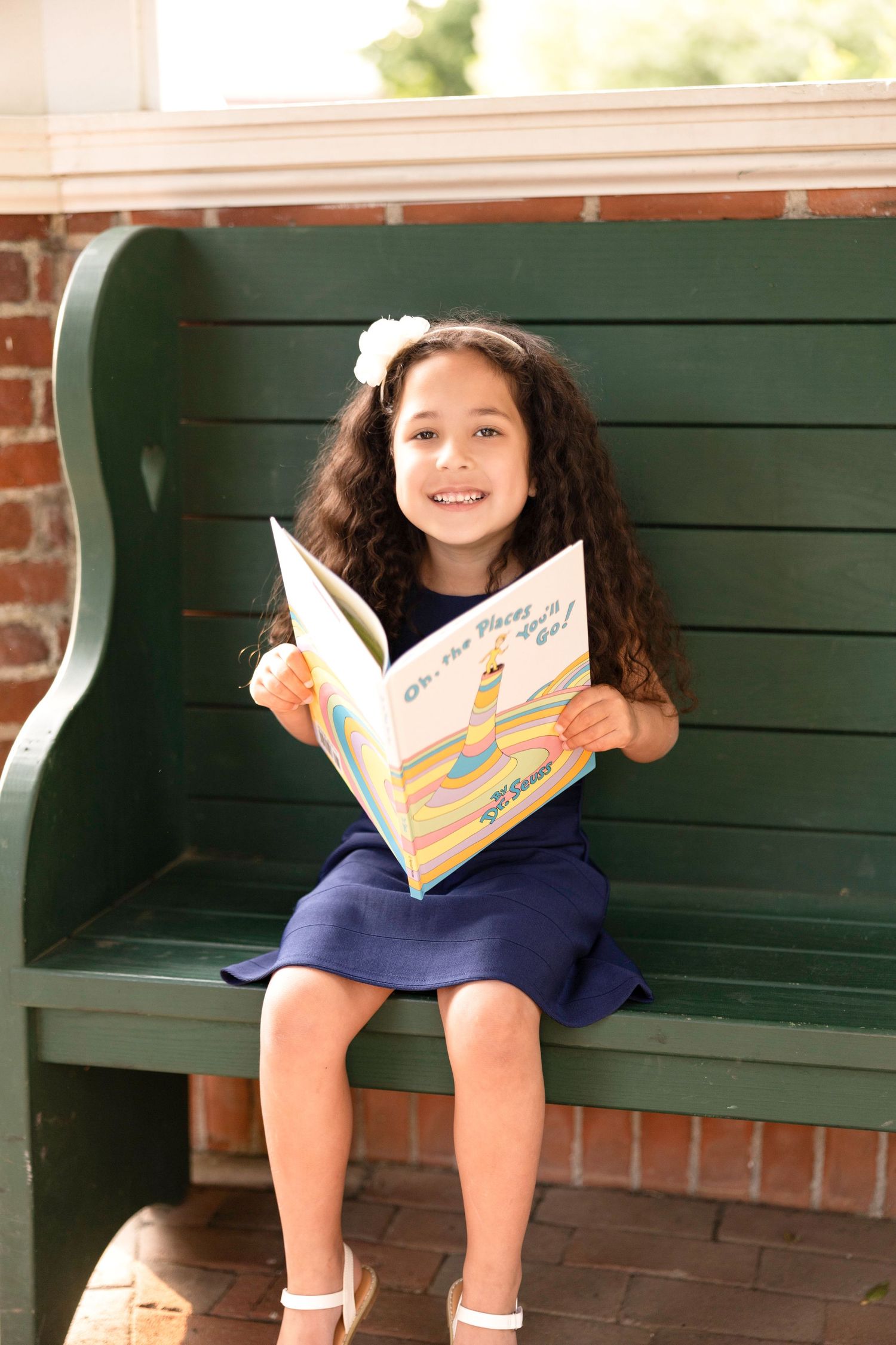 Child in navy dress reading book while sitting on green park bench.