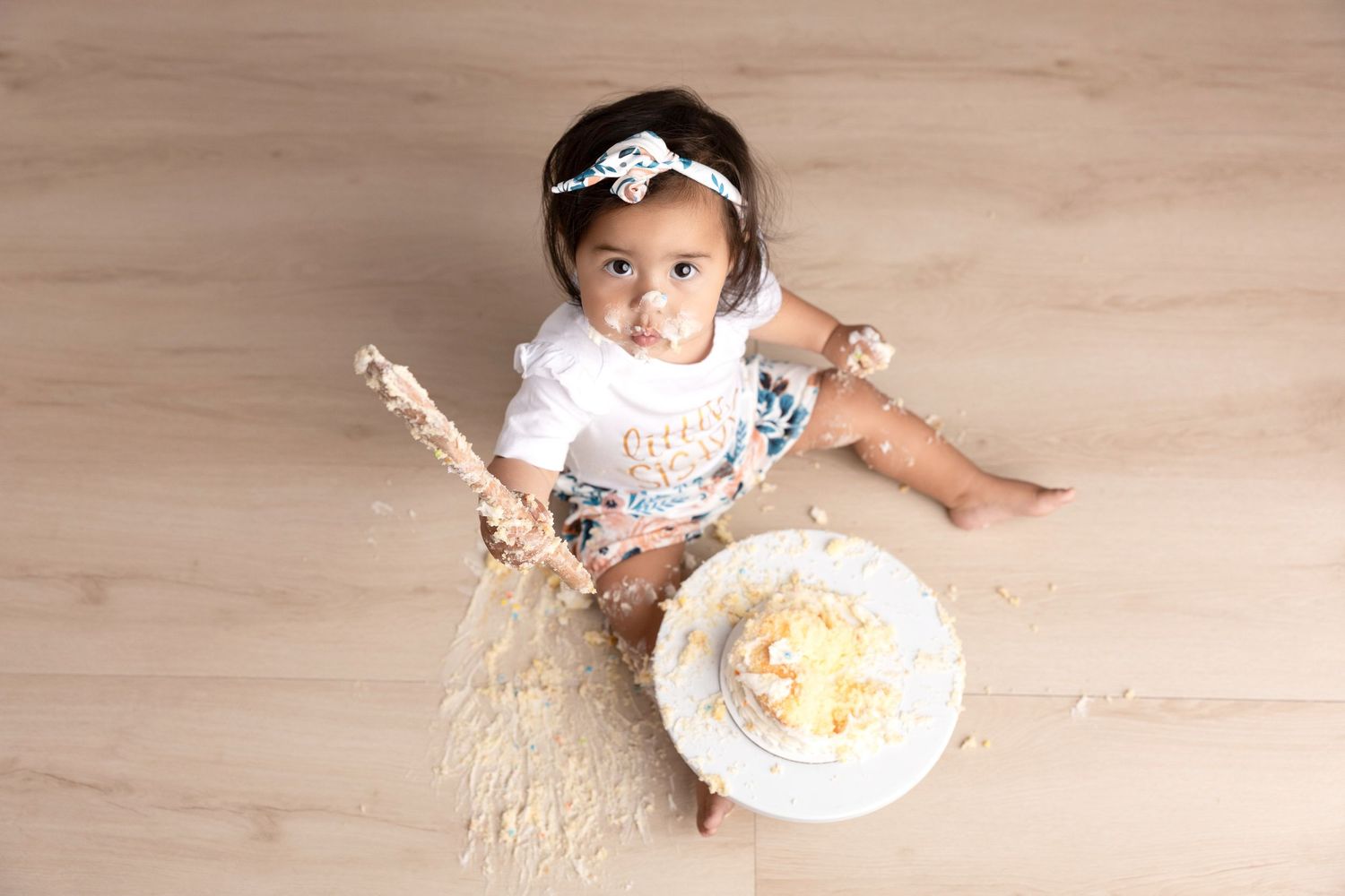 Baby's first birthday cake smash session on wooden floor with scattered frosting.