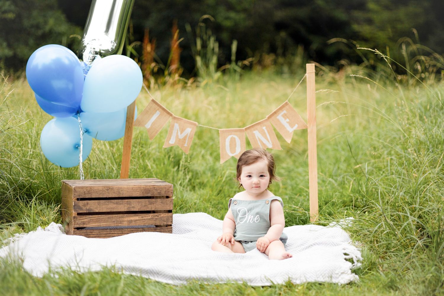 Baby's first birthday setup with wooden crate, blue balloons and white blanket in outdoor setting.