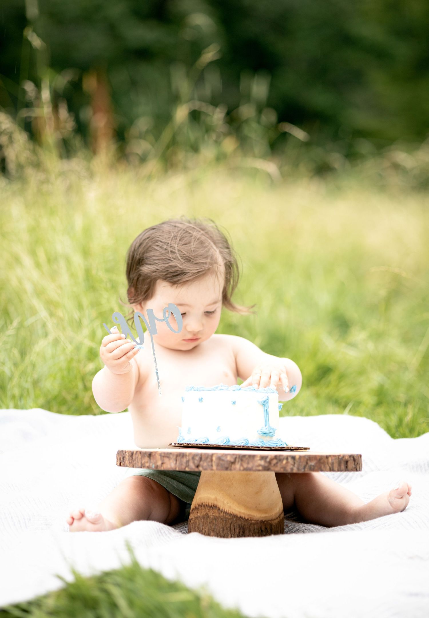 Baby exploring first birthday cake on rustic wooden slice outdoors.