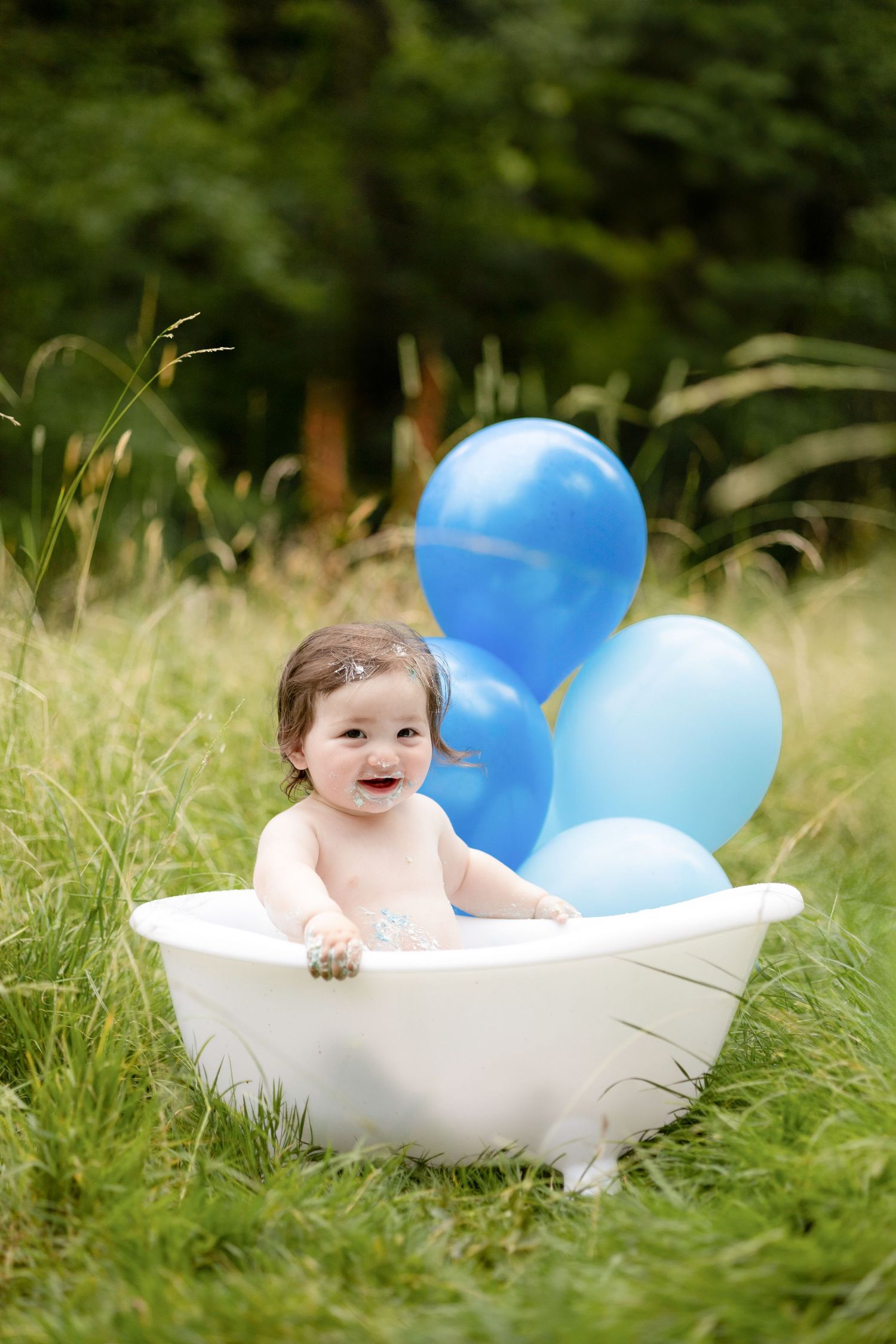 Baby taking a bath outdoors in a white tub surrounded by blue balloons and grass.