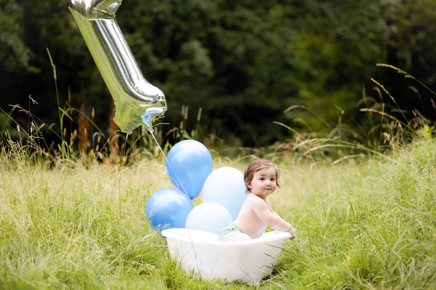 Baby bath photography setup in grassy field with white tub and blue balloons.