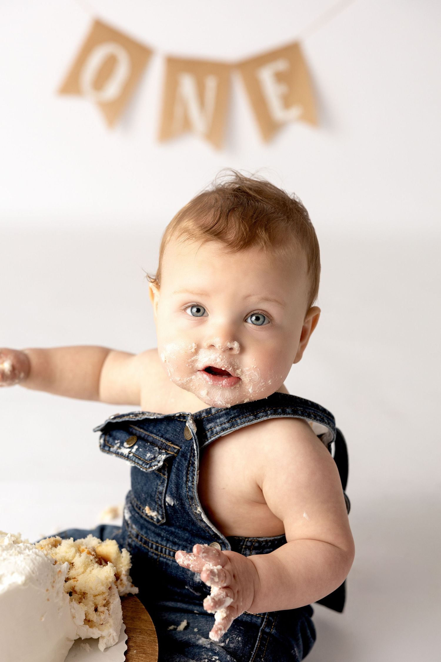 Baby in denim overalls enjoying messy cake smash during first birthday photoshoot.