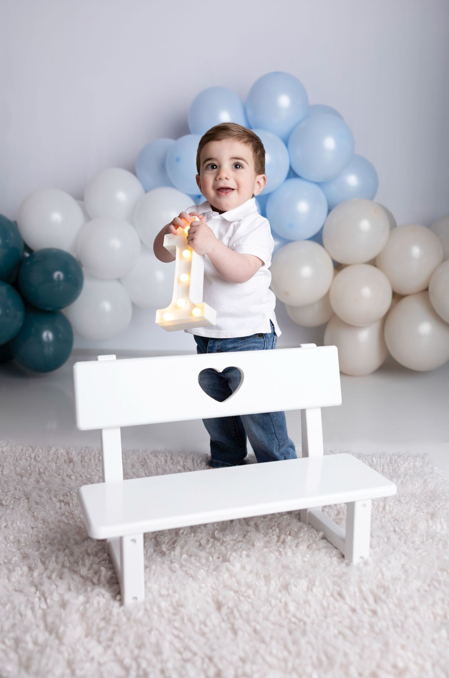 First birthday photoshoot with baby on white bench surrounded by blue and white balloons.