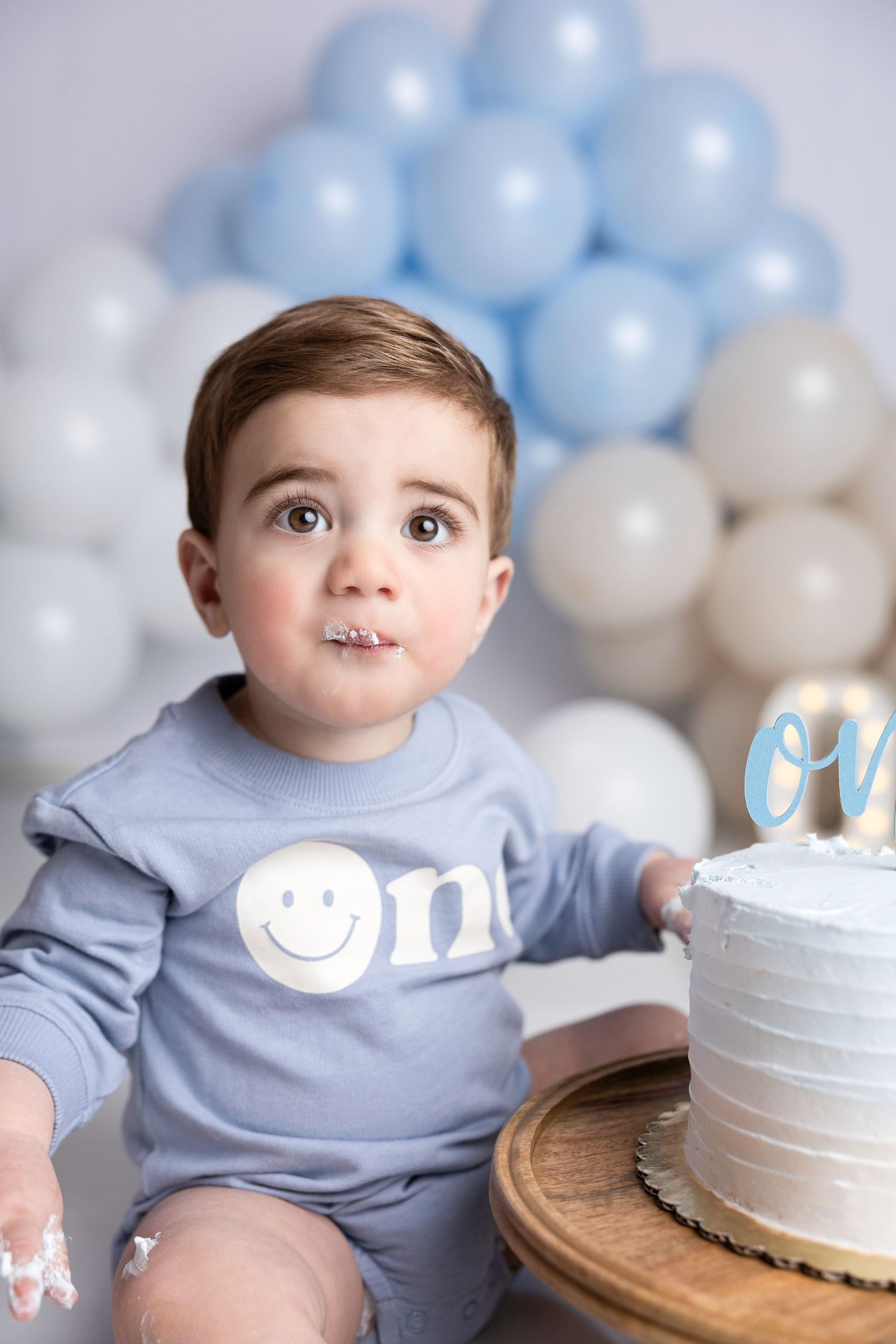 Baby in blue shirt sits by white birthday cake with balloon decorations.