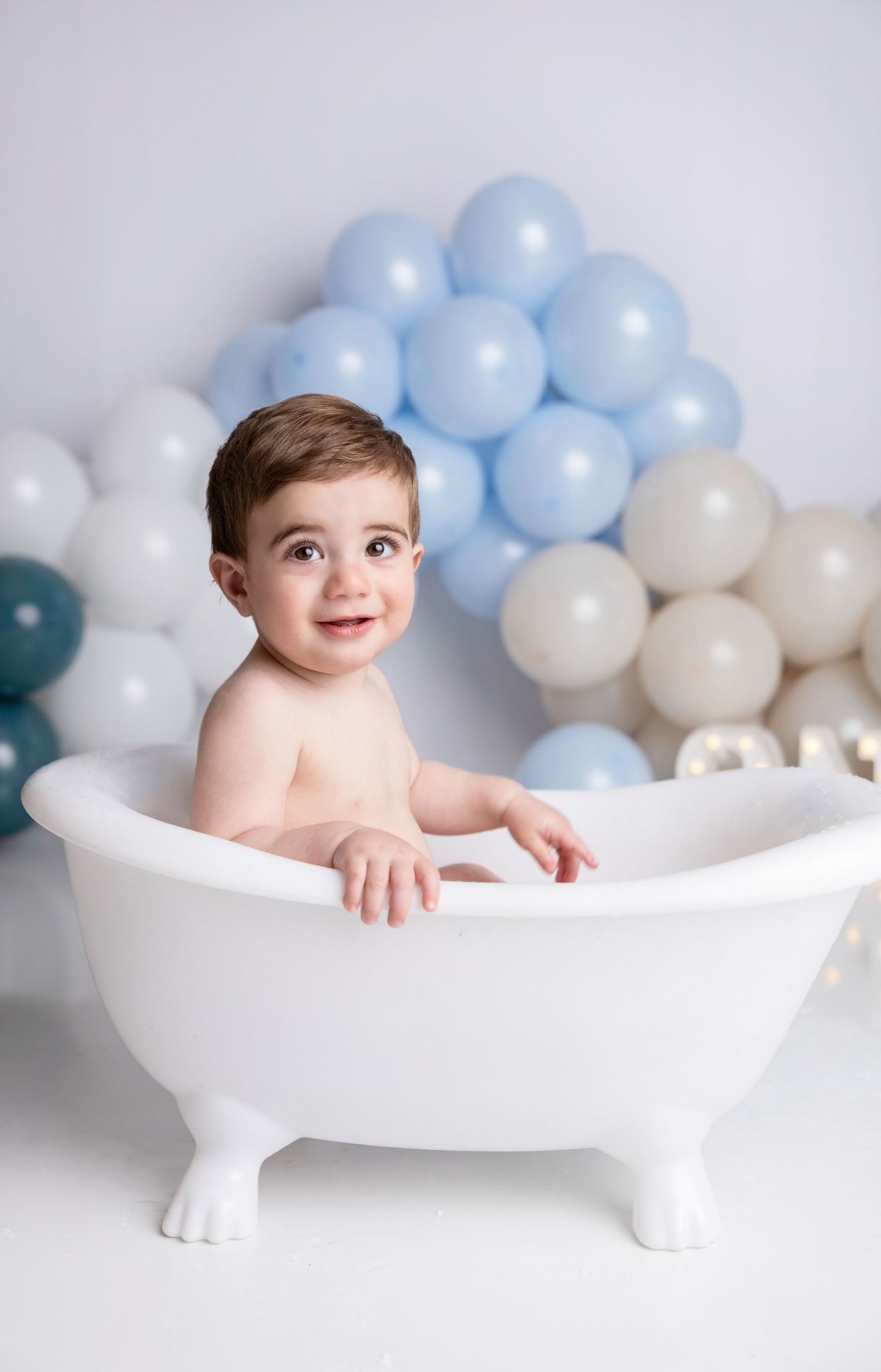 Baby sits in white vintage bathtub surrounded by soft blue and white balloons for milestone photo session.