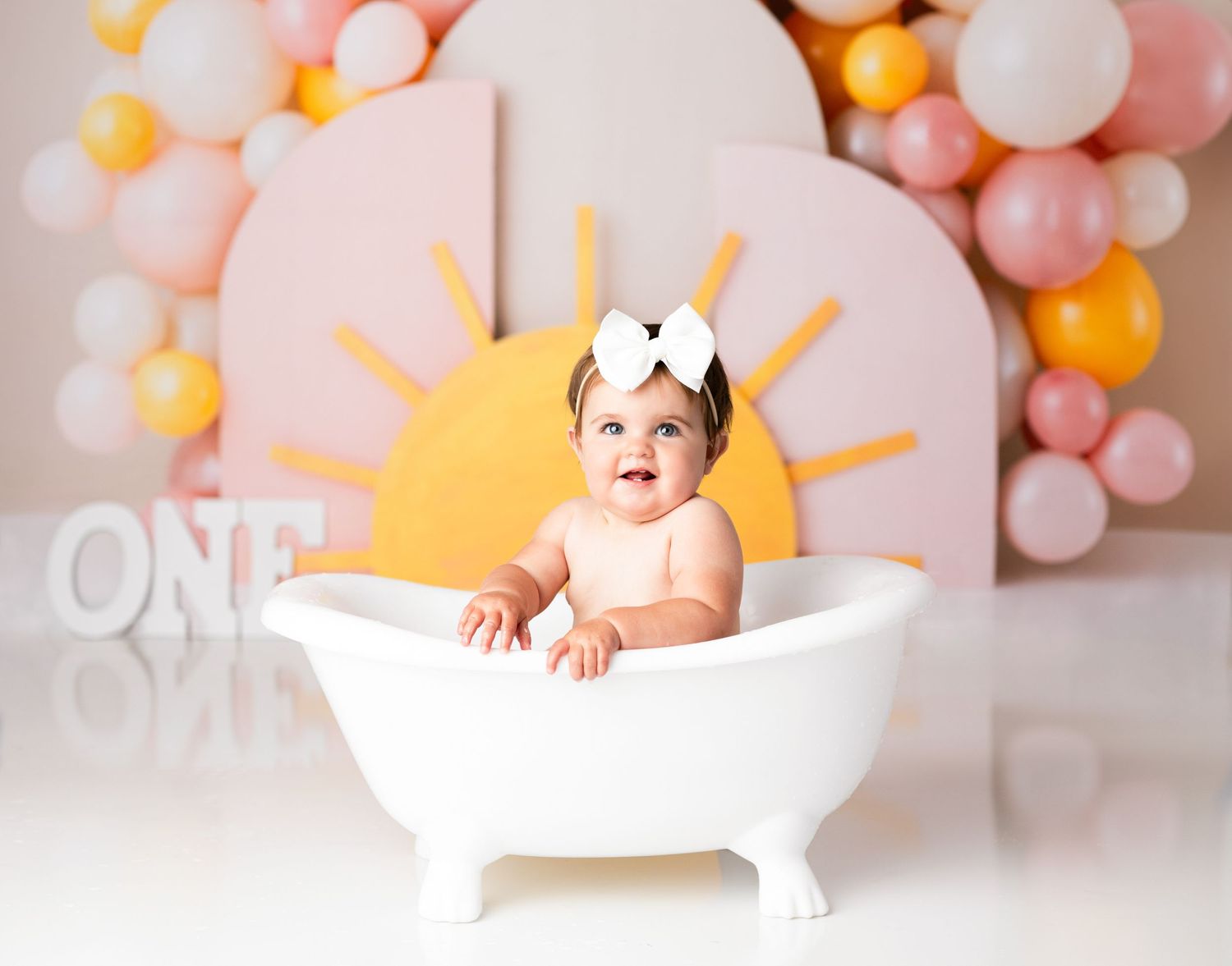 Baby sitting in white bathtub prop for first birthday photoshoot with balloon decorations.