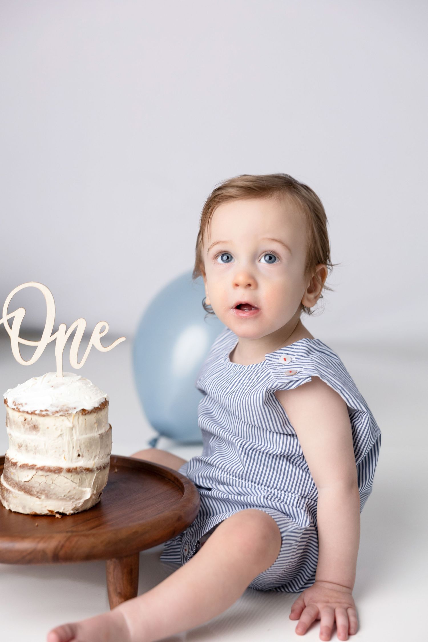 Baby in striped outfit sitting near small white cake and blue balloon.