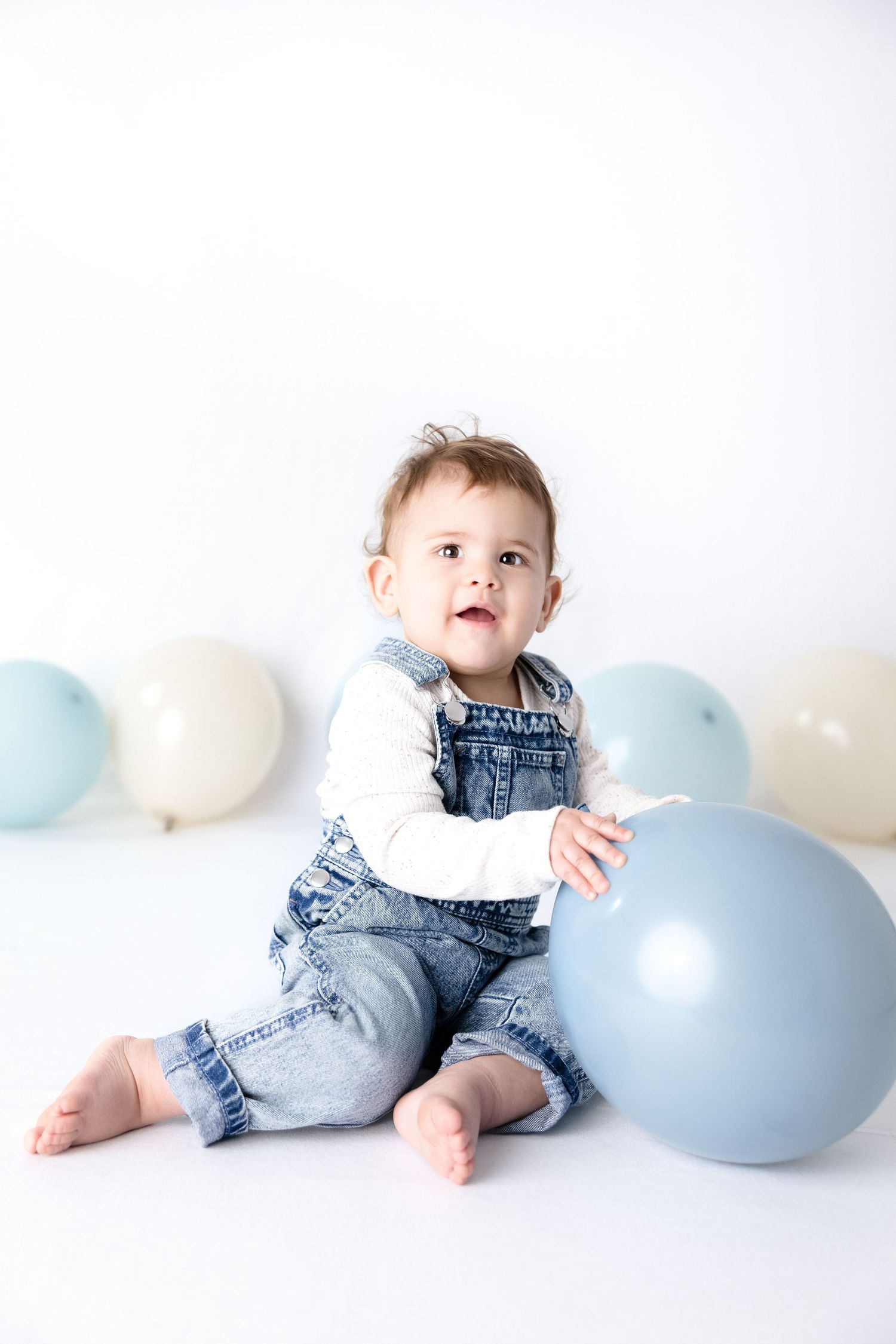 Baby in denim overalls plays with pastel blue and white balloons against white background.