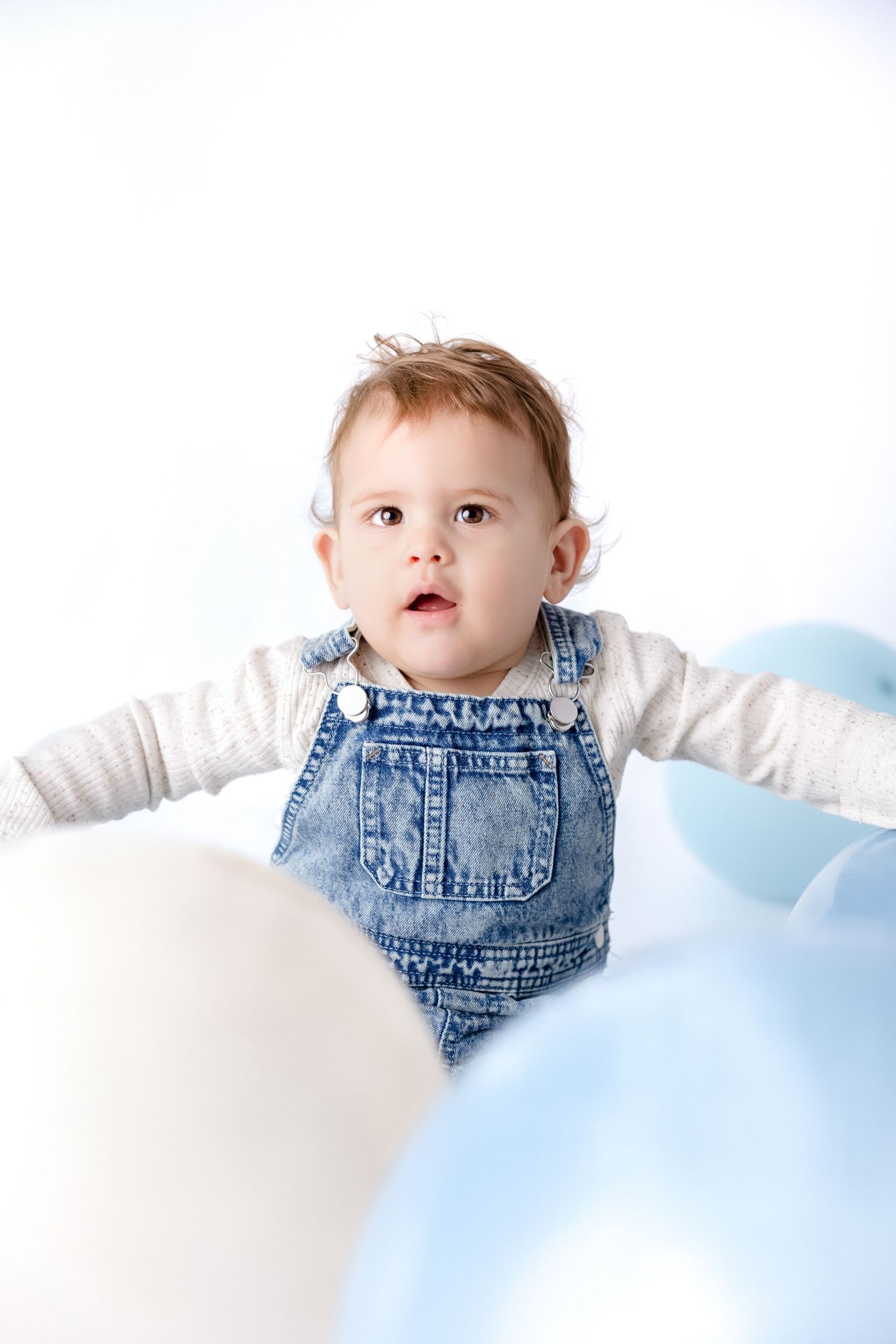 Baby wearing denim overalls lies on white surface with soft blue balloons.
