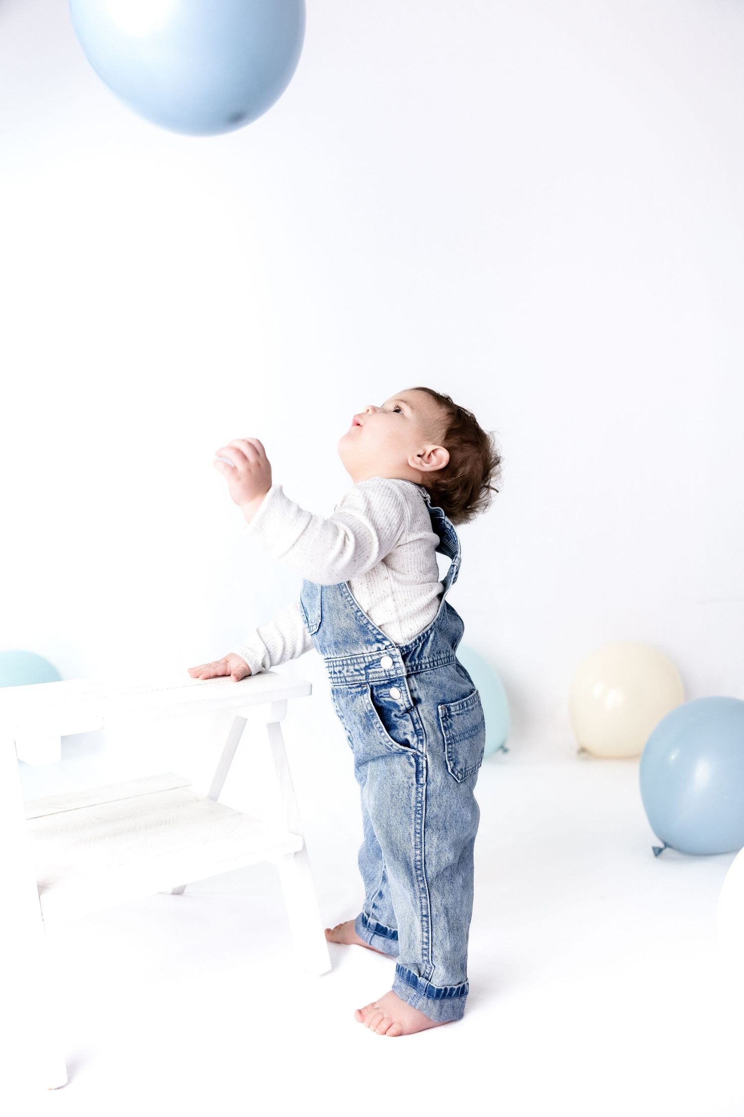 Baby in denim overalls stands and reaches for blue balloons in minimalist setting.